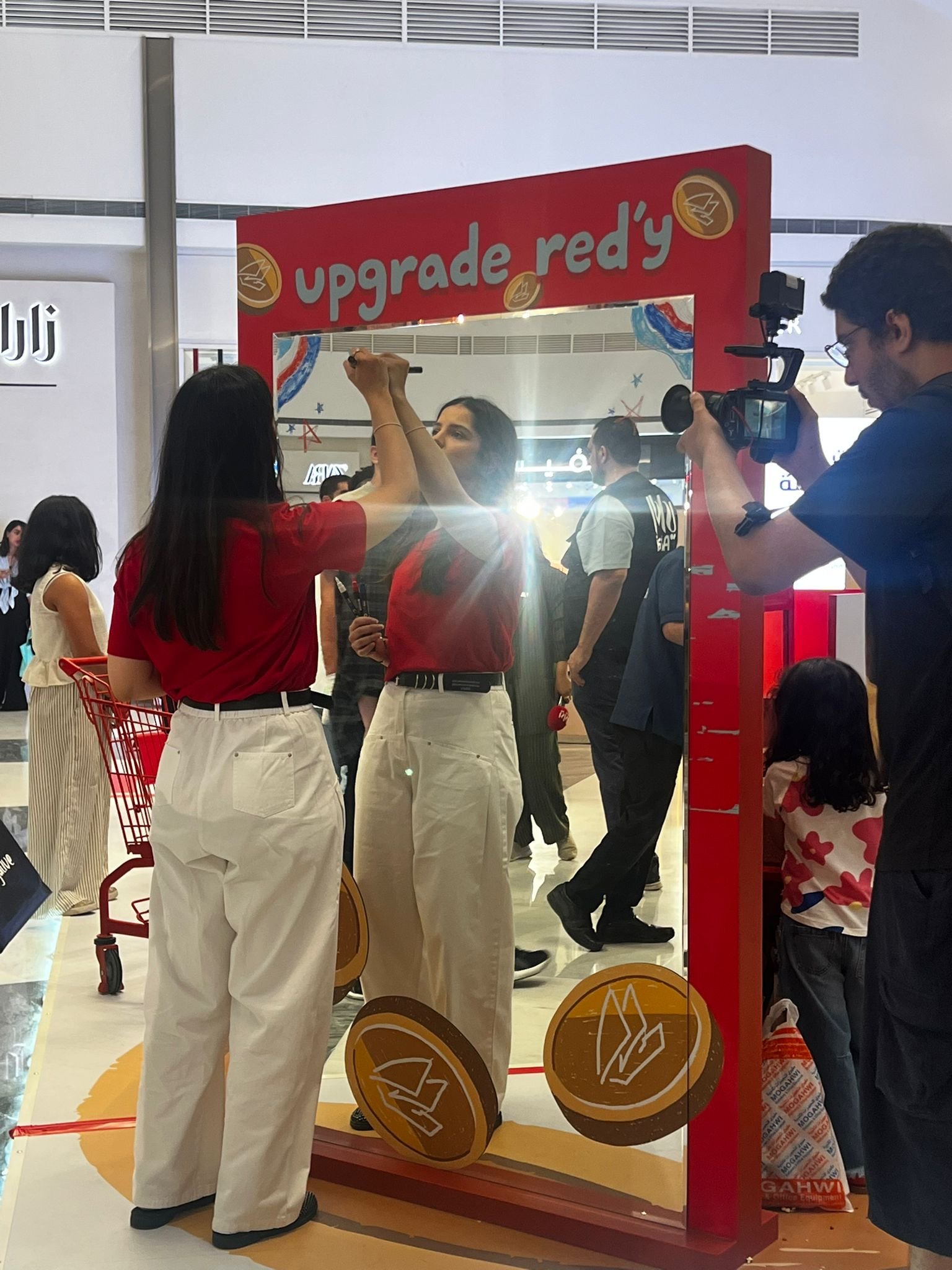 People gathered around a red promotional display labeled 'upgrade red'y' in a shopping mall. Some individuals are participating in a promotional activity, with a woman signing a mirror and a man recording with a camera. The display has gold coins with a logo and appears to be part of a marketing event.