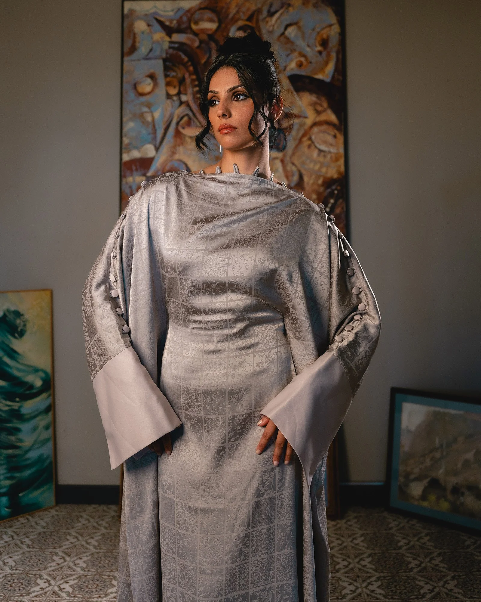 A woman with dark hair in an elegant updo, wearing a silver embroidered dress with bell sleeves, standing in an art gallery with paintings in the background.