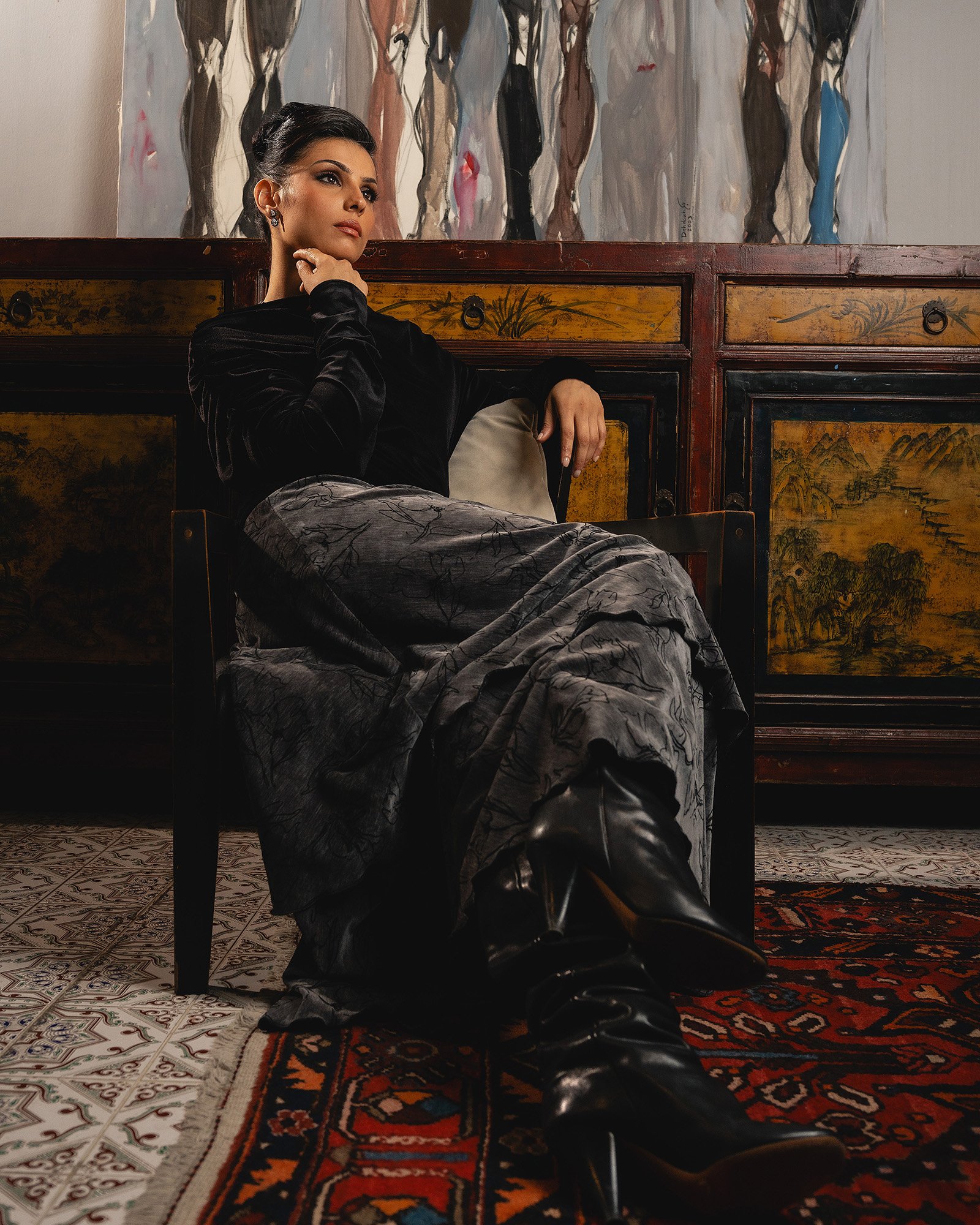 A woman with dark hair and makeup sitting on a chair in a room with patterned rugs and an embroidered Asian-style cabinet. She is wearing a dark velvet top, patterned pants, and black boots, with her right hand touching her face and her left arm rest