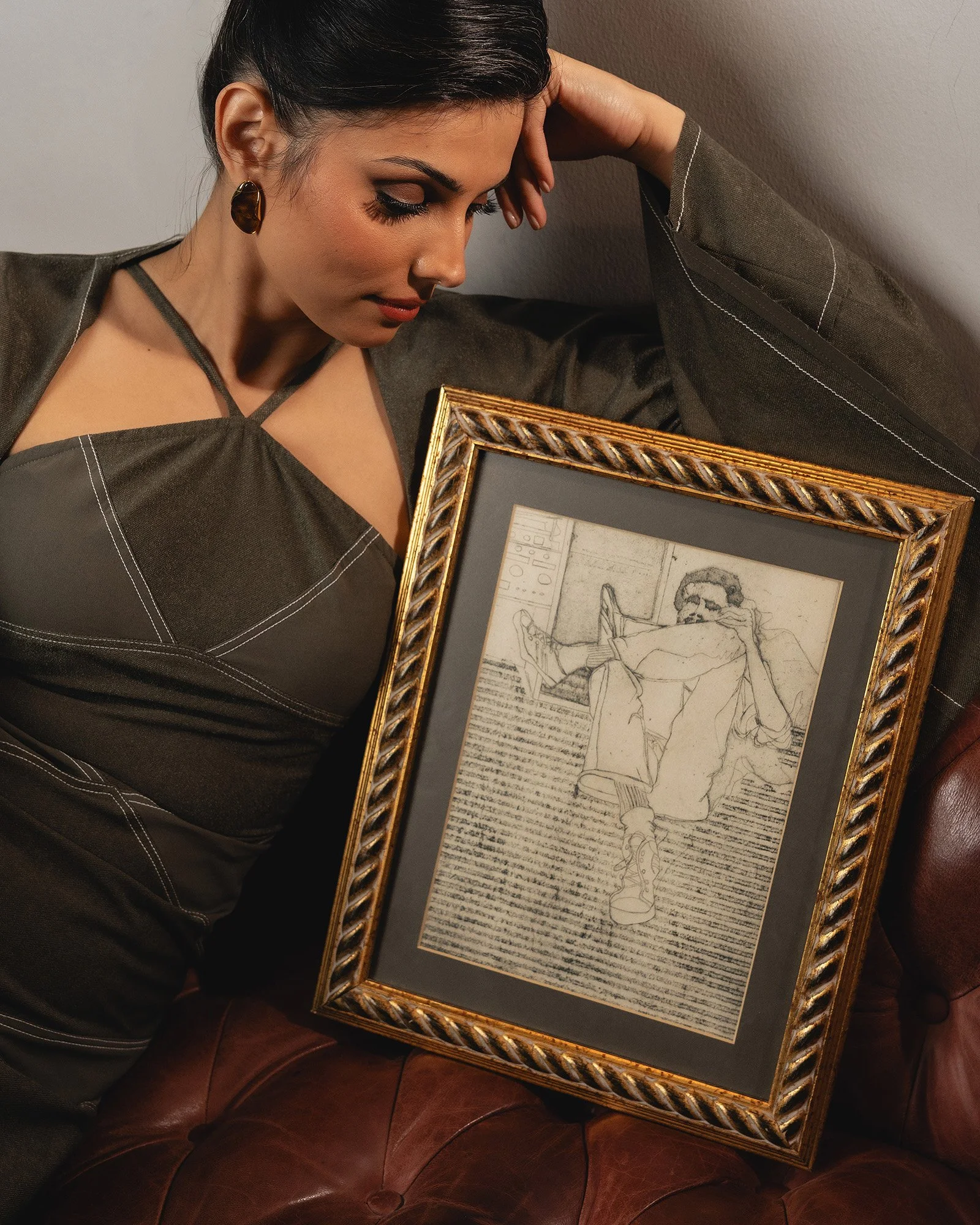 A woman with dark hair and earrings, wearing a dark brown top, is resting her head against her hand while looking down at a framed sketch of a person seated with their legs crossed and holding what appears to be a phone, placed on a brown leather cou