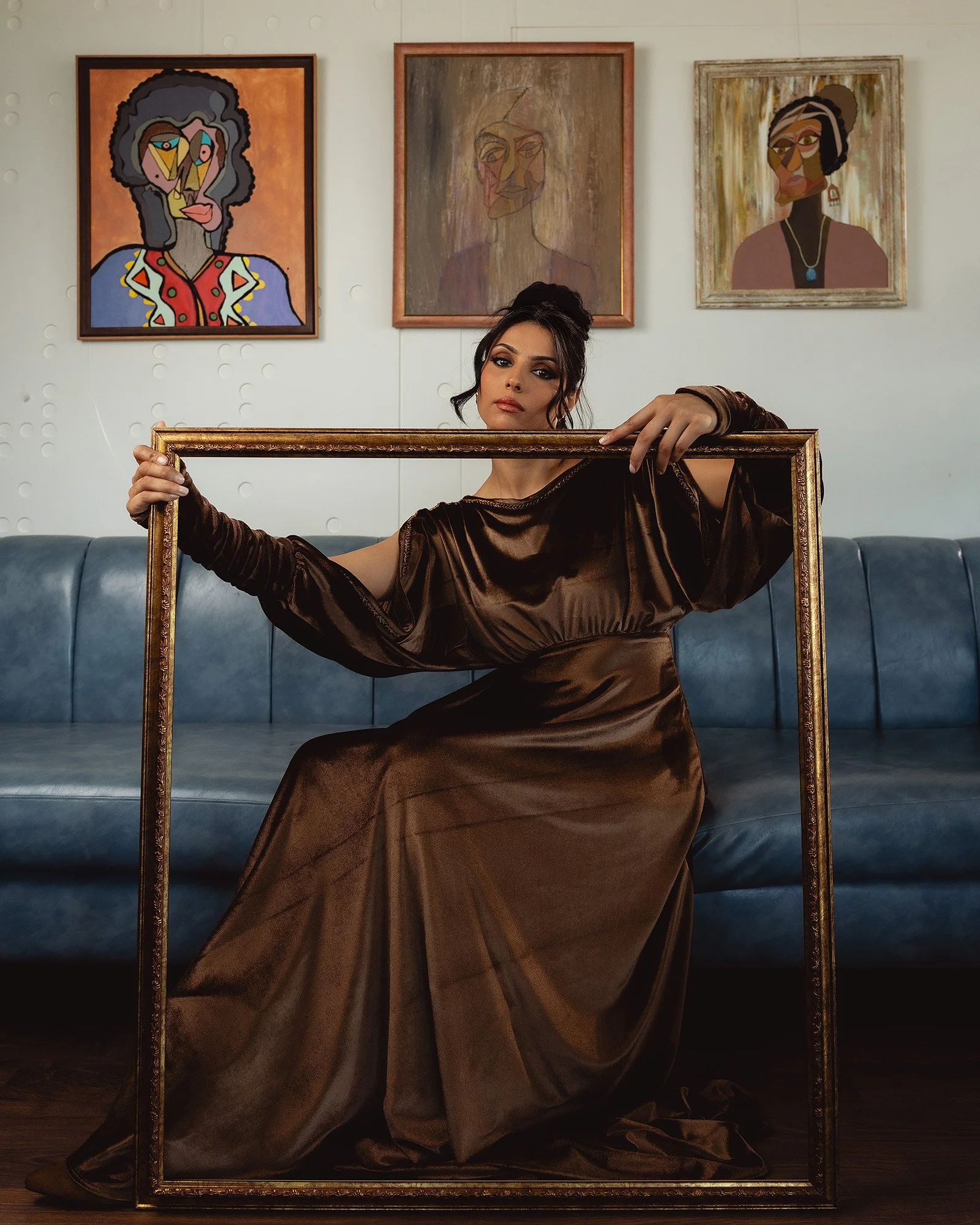 A woman in a shiny brown dress sits on a blue couch, holding an empty ornate gold frame in front of her. Behind her, there are three framed art portraits on a white wall. The woman has dark hair styled in an updo and is looking slightly to the side.
