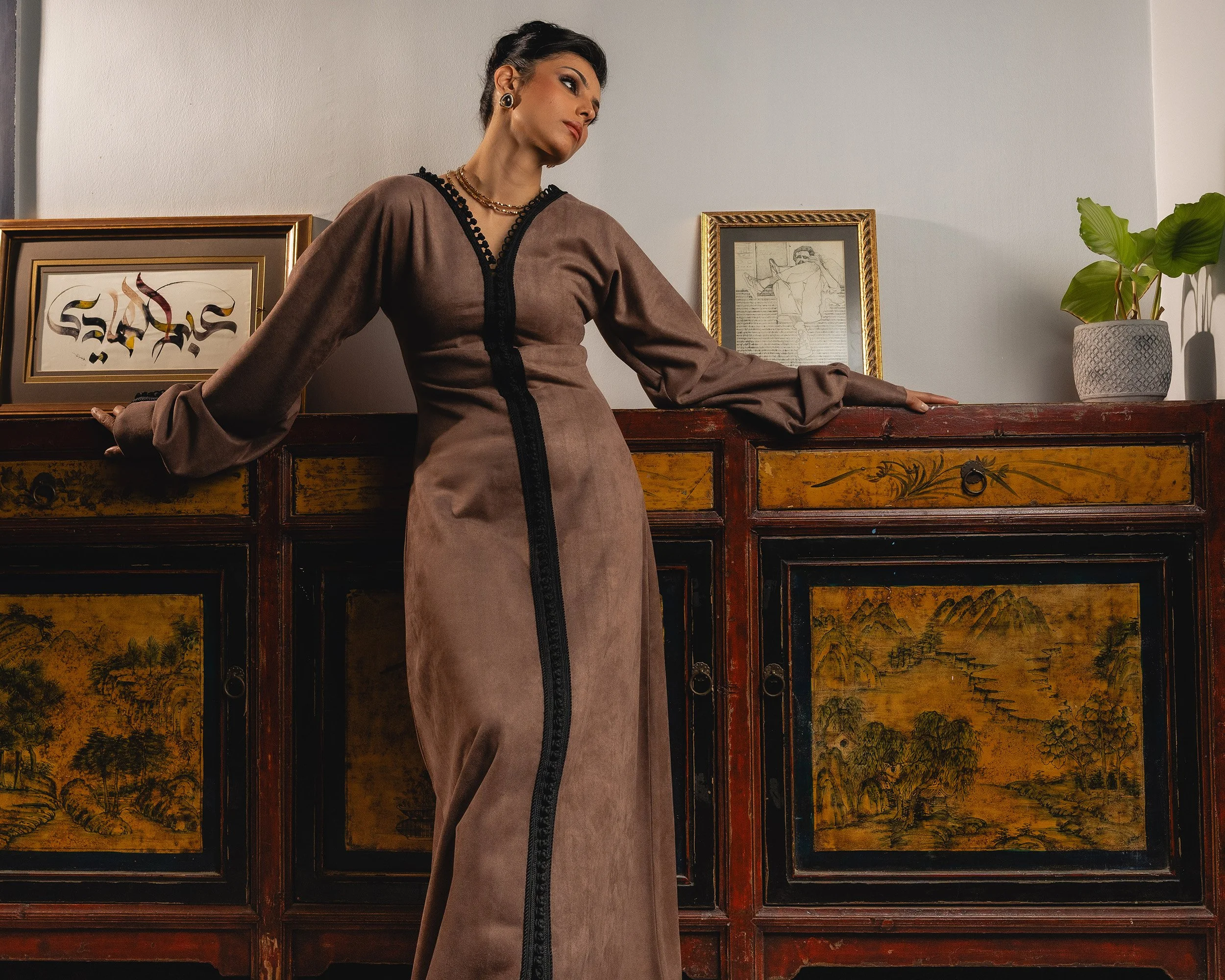 A woman with dark hair styled up, wearing a brown dress with black trim and jewelry, leaning on a vintage wooden chest with artwork and a potted plant in the background.