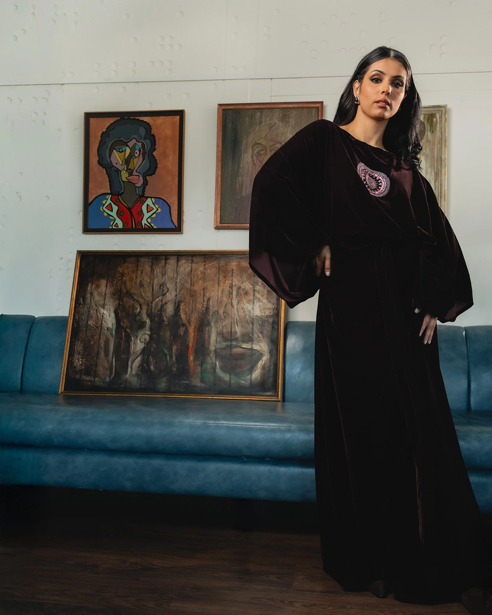 A woman with dark hair and makeup, wearing a dark velvet dress with embroidered details, standing in an art gallery. Behind her, there are three framed paintings leaning against the wall, including a colorful abstract portrait and a muted-toned lands