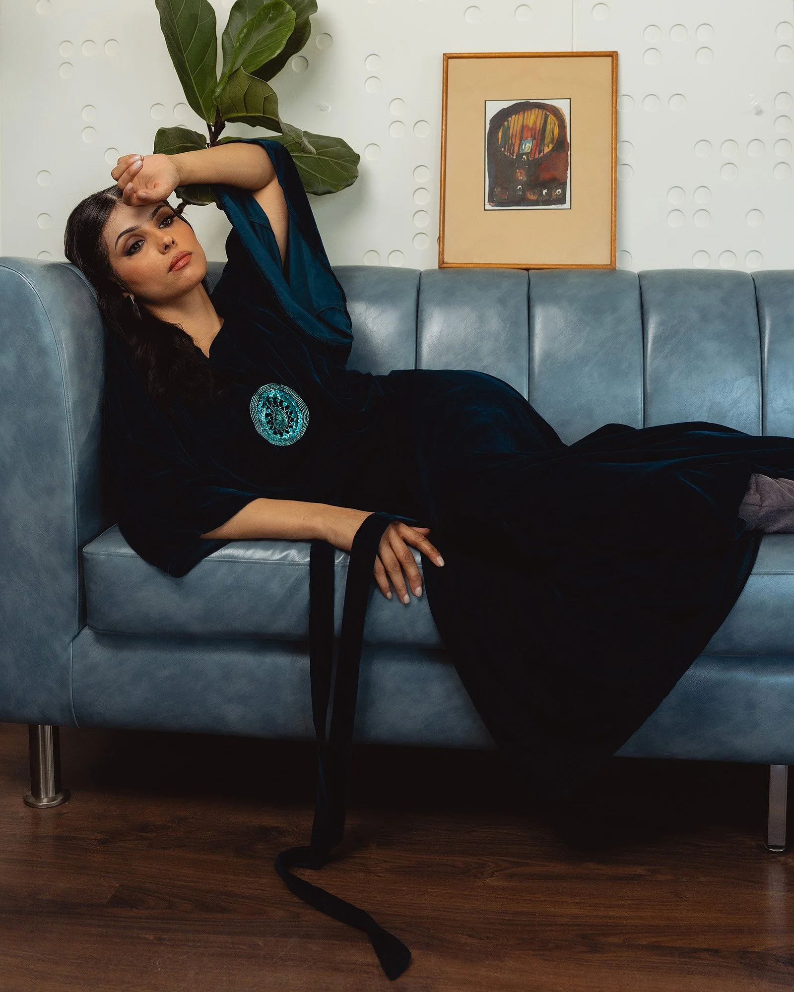 A woman with dark hair and makeup reclining on a blue couch, holding her forehead with her right arm. She wears a dark velvet dress with an embroidered circular blue and black patch on the chest, and has a black strap hanging from her wrist. Behind h