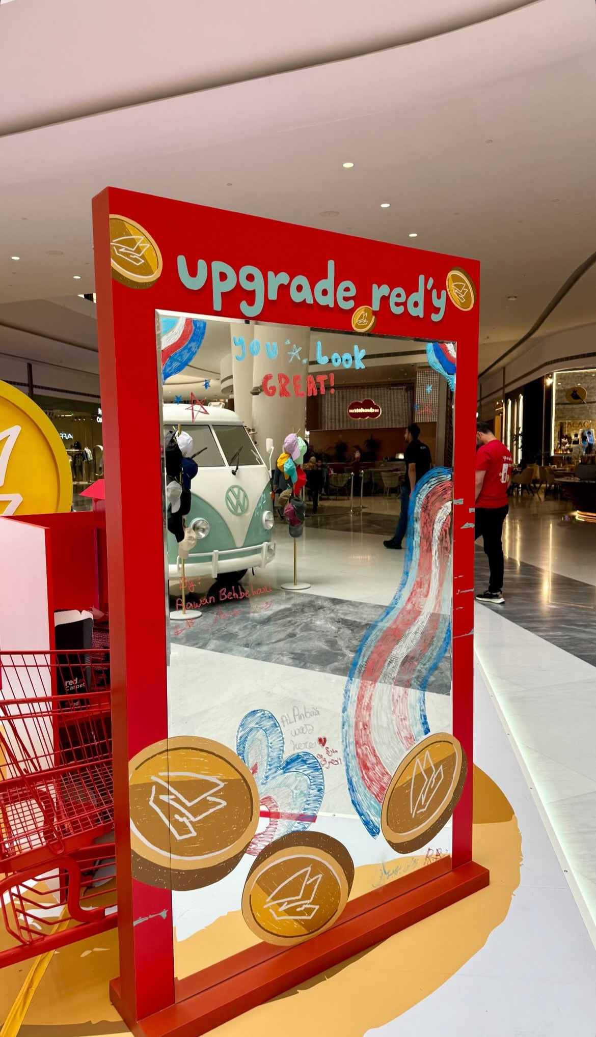 A red and yellow promotional display with a mirror in a shopping mall. The display reads 'upgrade red' and has musical notes and dollar signs on it. The mirror reflects a Volkswagen van, shopping carts, and mall patrons.