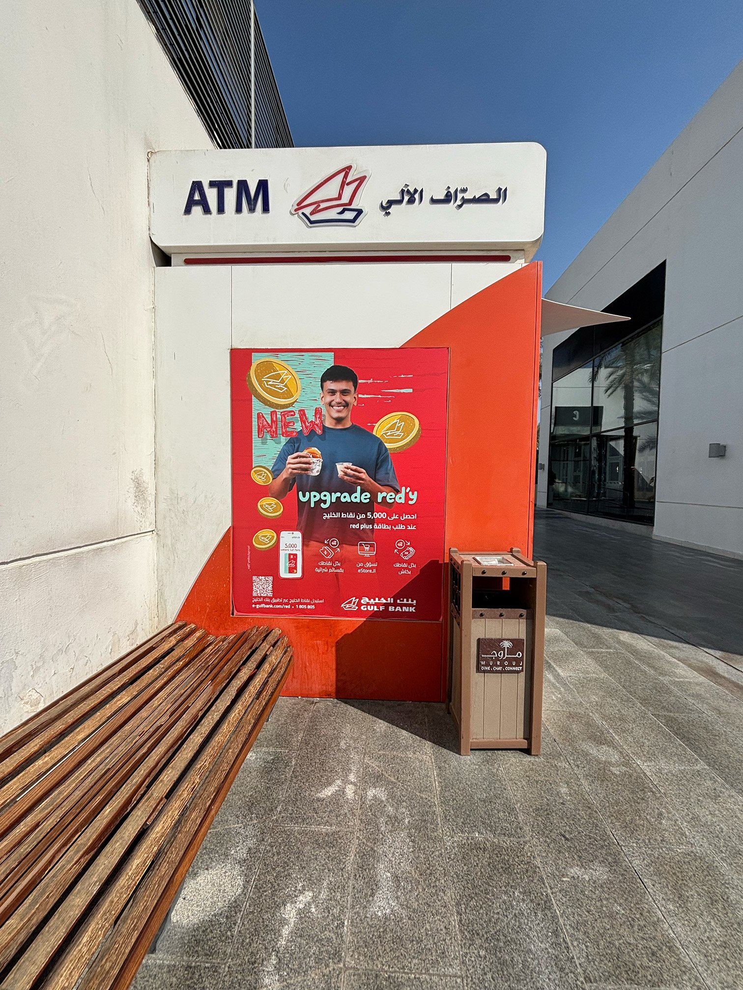 A sign with Gulf Bank logo and text in Arabic and English, advertising for a new promotion to upgrade to red card plus for earning 5,000 points. In the background, a modern building with glass windows and a palm tree reflection, and a wooden bench on a tiled pavement.