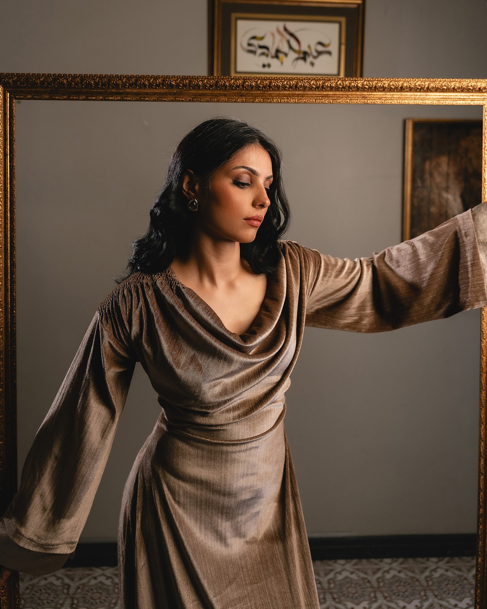 A woman with black hair in a brown satin dress adorned with fringes, posing inside a gold frame mirror, with framed artwork on a neutral wall in the background.