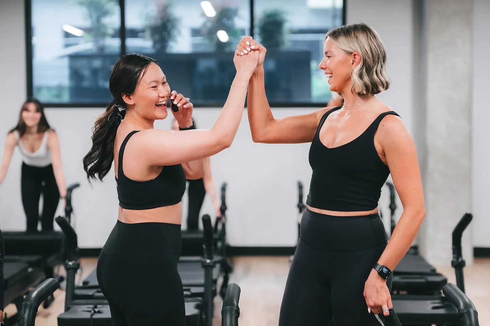 Two women in fitness attire high-fiving each other in a gym with treadmills, with two more women in workout clothes in the background.