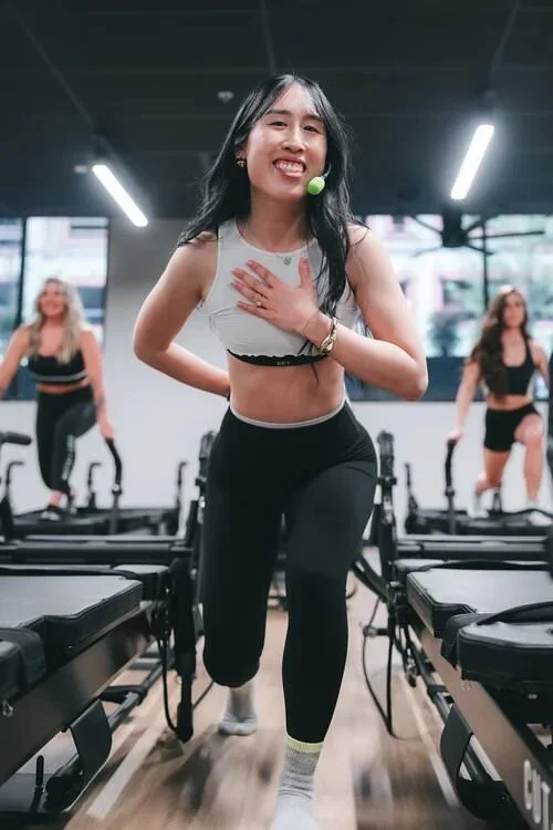 A woman leading a fitness class in a gym, smiling with a hand on her chest, wearing workout clothes, and using a microphone. Two other women are in the background exercising on reformer machines.