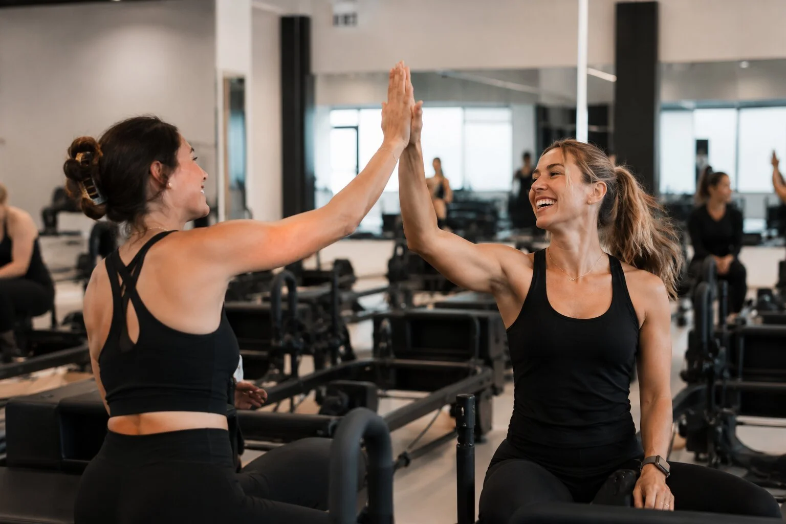 Two women in black workout clothes are giving each other a high five in a fitness studio, with other people exercising in the background.