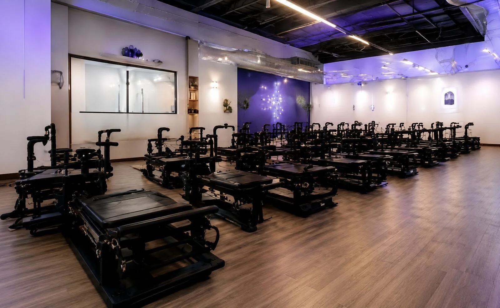 A Pilates or fitness studio room with multiple Black reformer machines arranged in rows on a wooden floor. The room has white and blue walls with minimal decorations and lighting fixtures, and a large window or mirror on one wall.