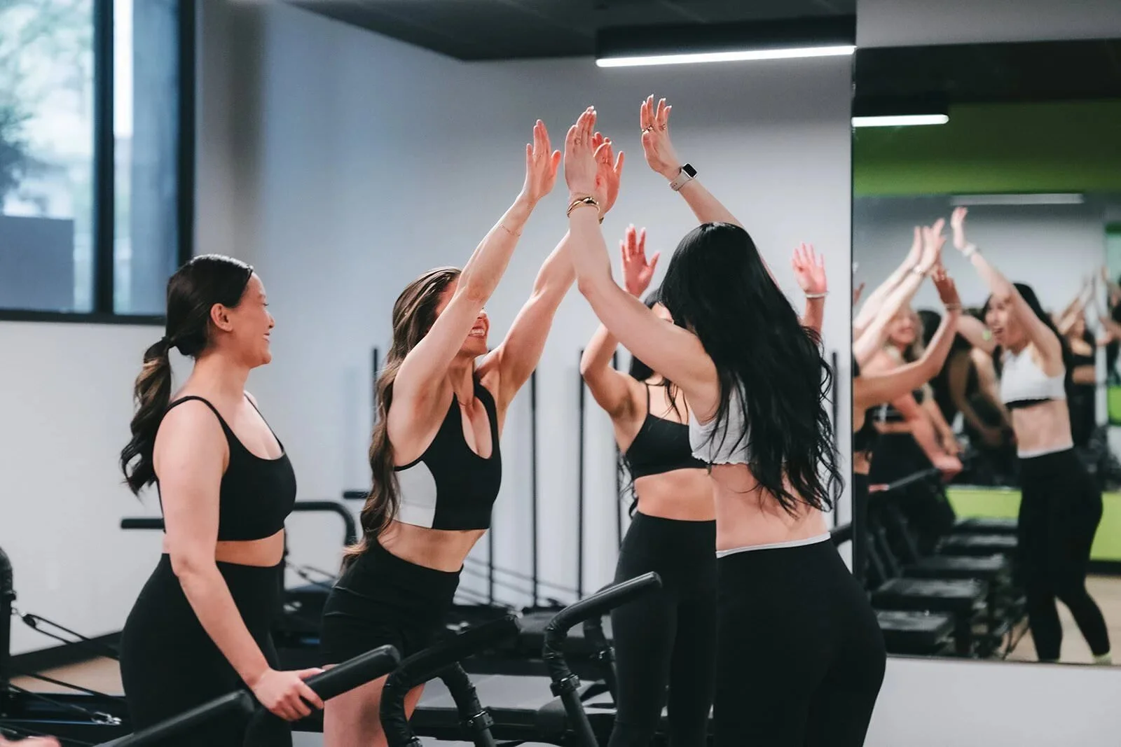 A group of women wearing workout clothes high-five in a fitness class at the gym, with a mirror reflecting their celebration.