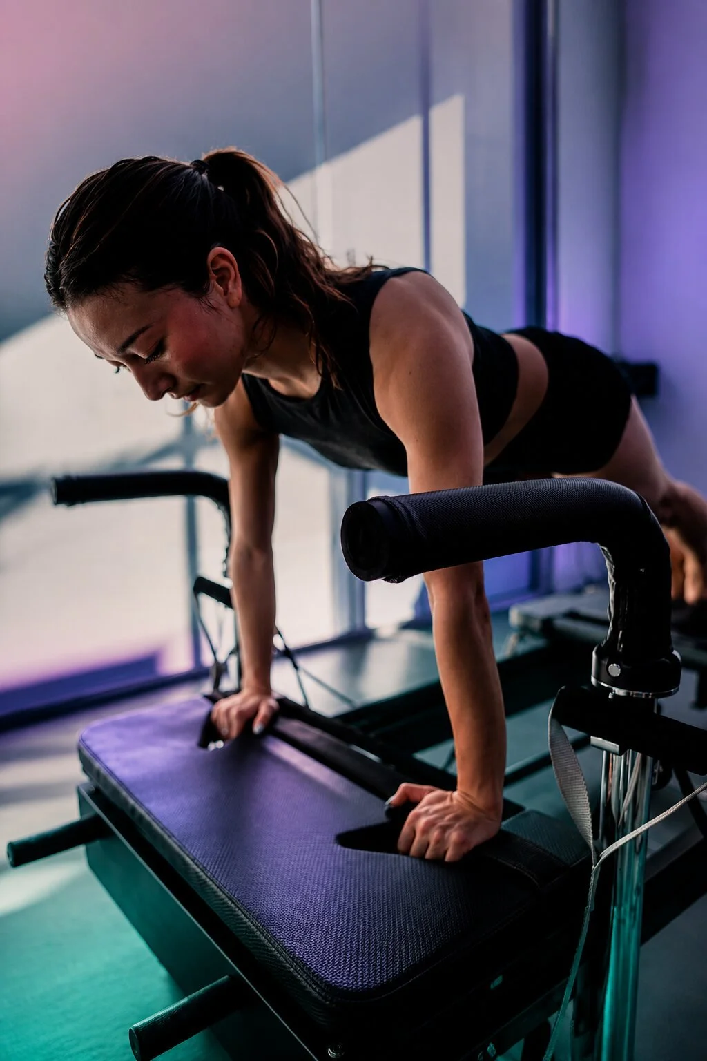 A woman doing a push-up on a reformer Pilates machine in a gym with large windows and colorful lighting.
