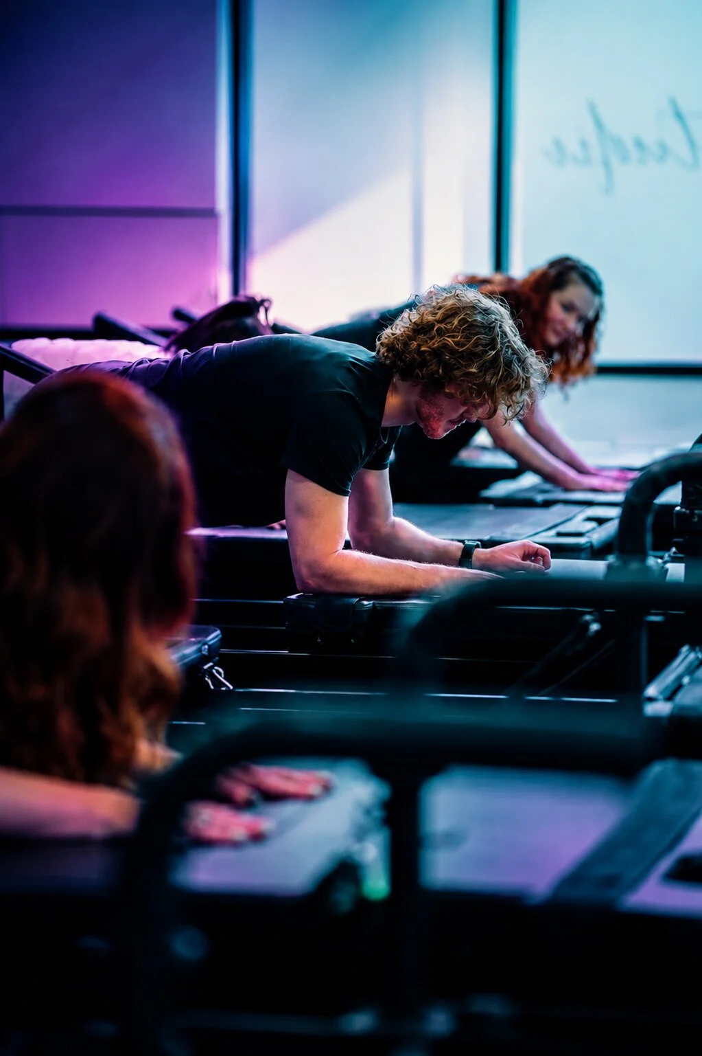 People participating in a fitness class, lying on their stomachs on mats, following an instructor in a dimly lit room with colorful lighting, and a whiteboard in the background.