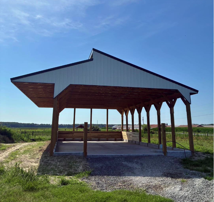 A wooden open-sided pavilion with a metal roof, situated on a gravel surface in a grassy field with a clear blue sky in the background.