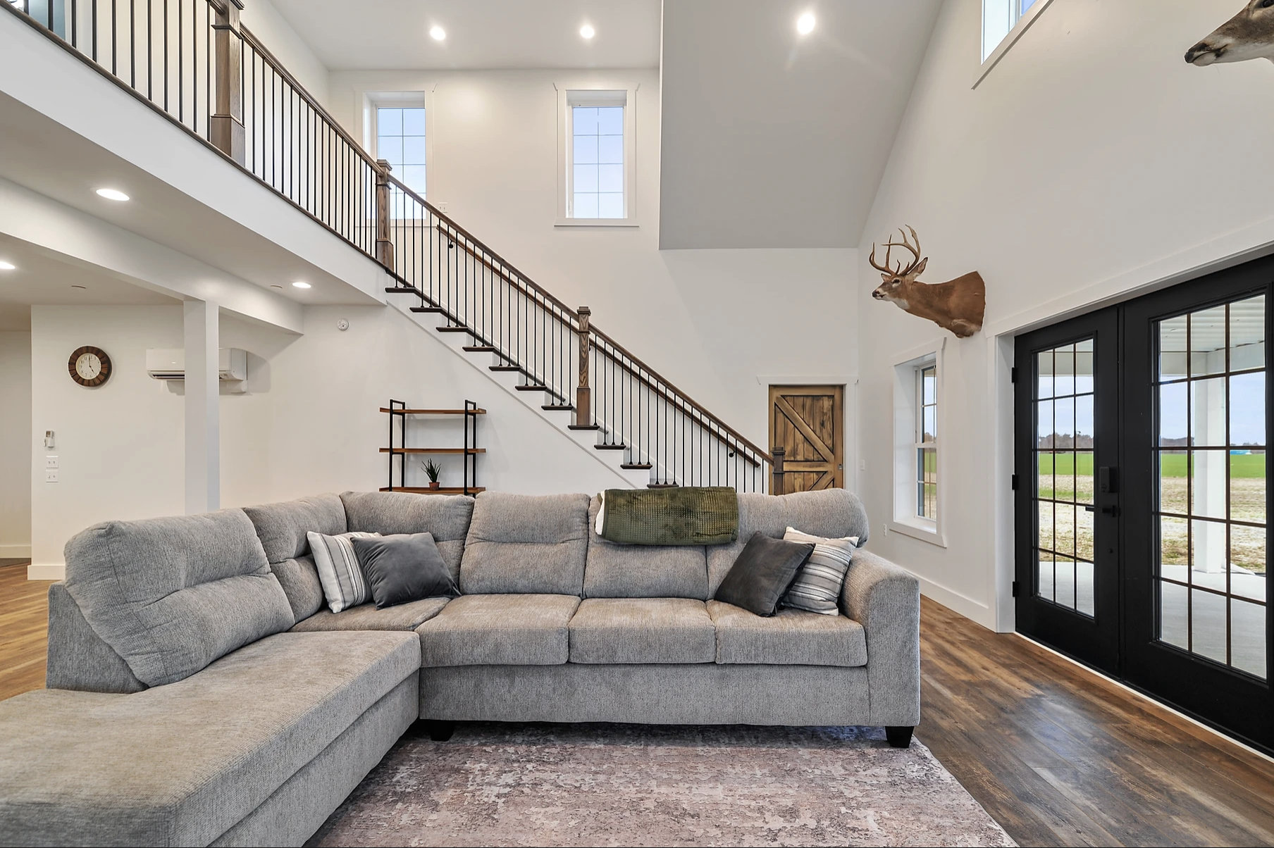 Living room with gray sectional sofa, decorative pillows, a green throw blanket, and a wooden side table with a small plant. There is a staircase with black metal railing and wooden handrail, two tall windows, and wooden door with black double glass doors leading outside. Mounted deer head visible on the wall.