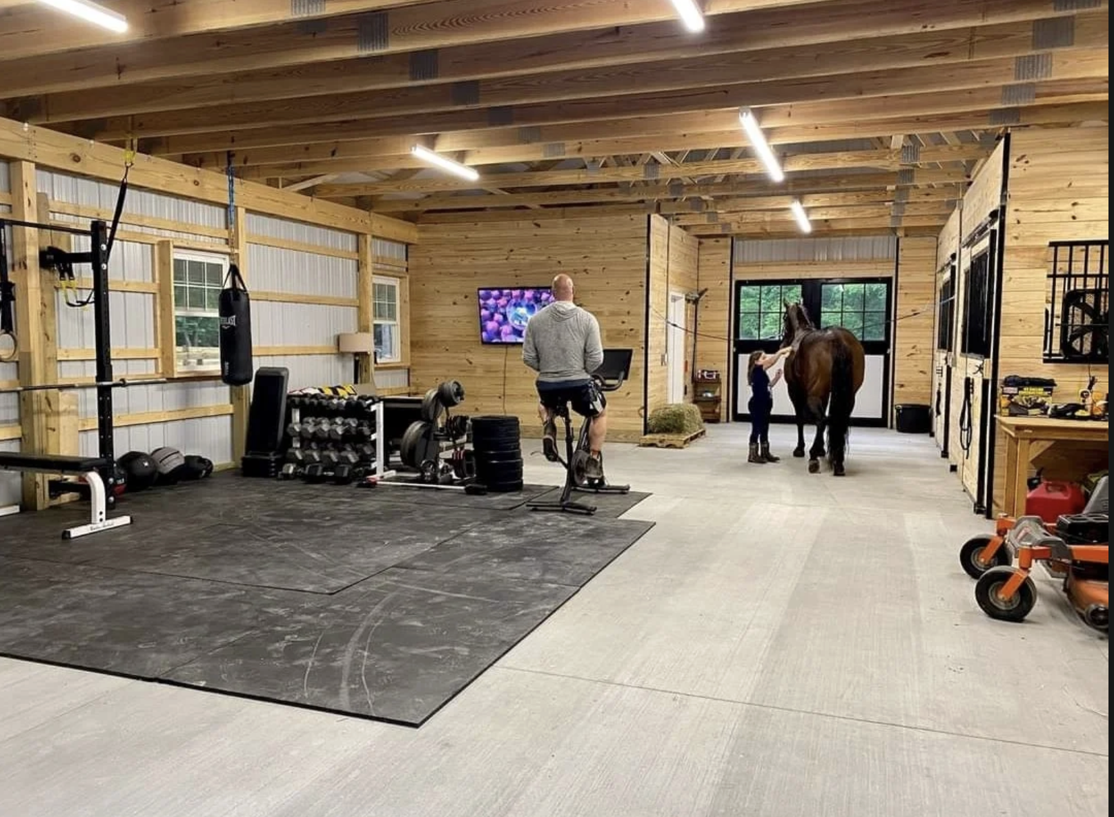 Interior of a wooden gym with fitness equipment, a man on a stationary bike, a person grooming a horse, and various workout and horse tack accessories.
