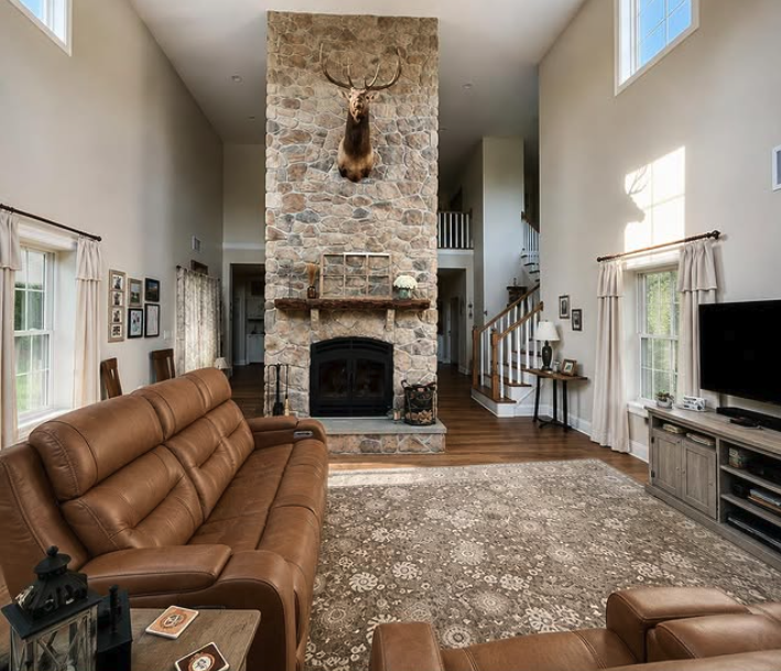 Living room with a stone fireplace and mounted deer head, brown leather sofa, TV on a stand, and windows with curtains.