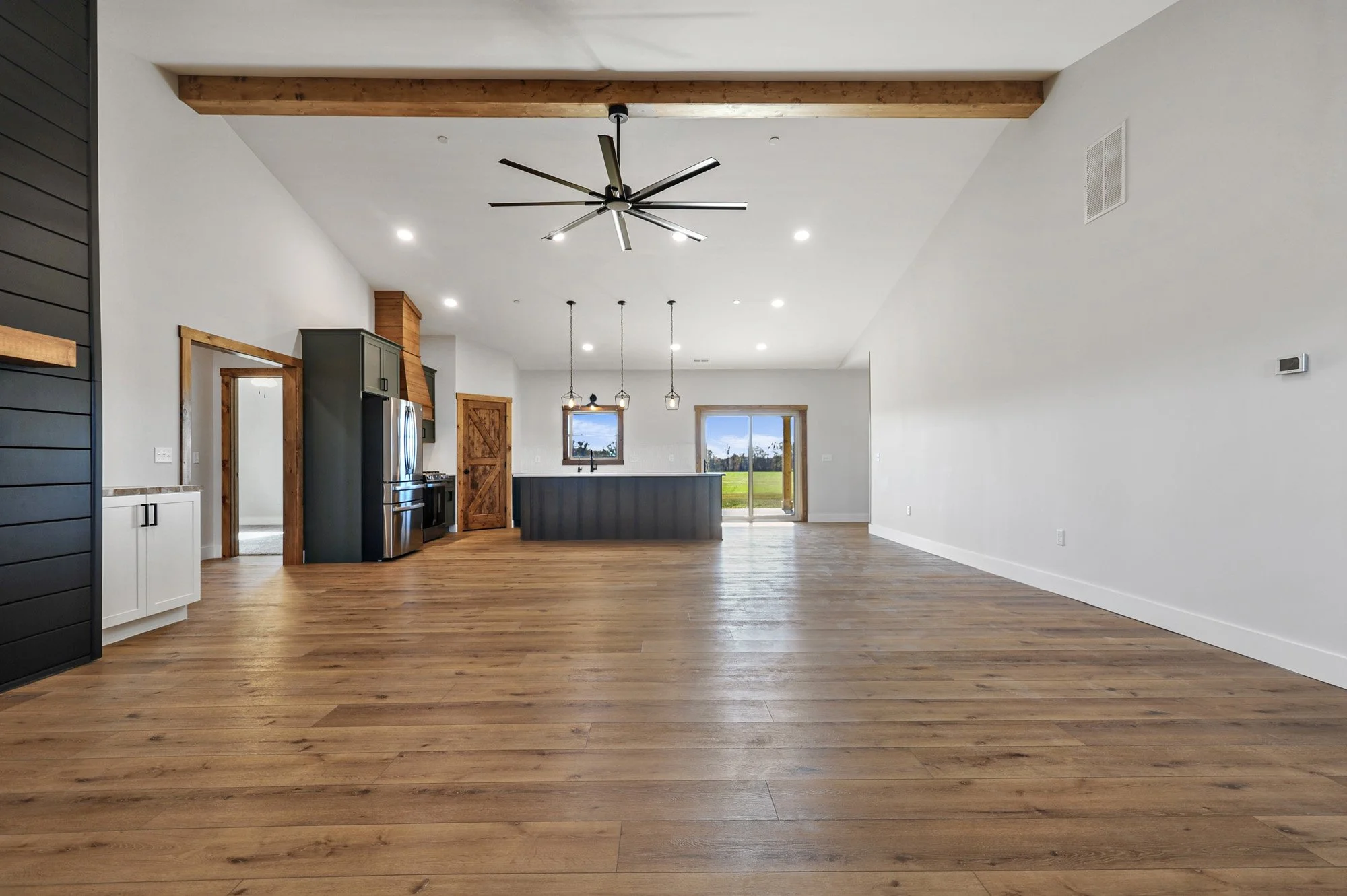 Open concept living space with a kitchen in the background, wooden flooring, a ceiling fan, and large windows showing a scenic outdoor view.