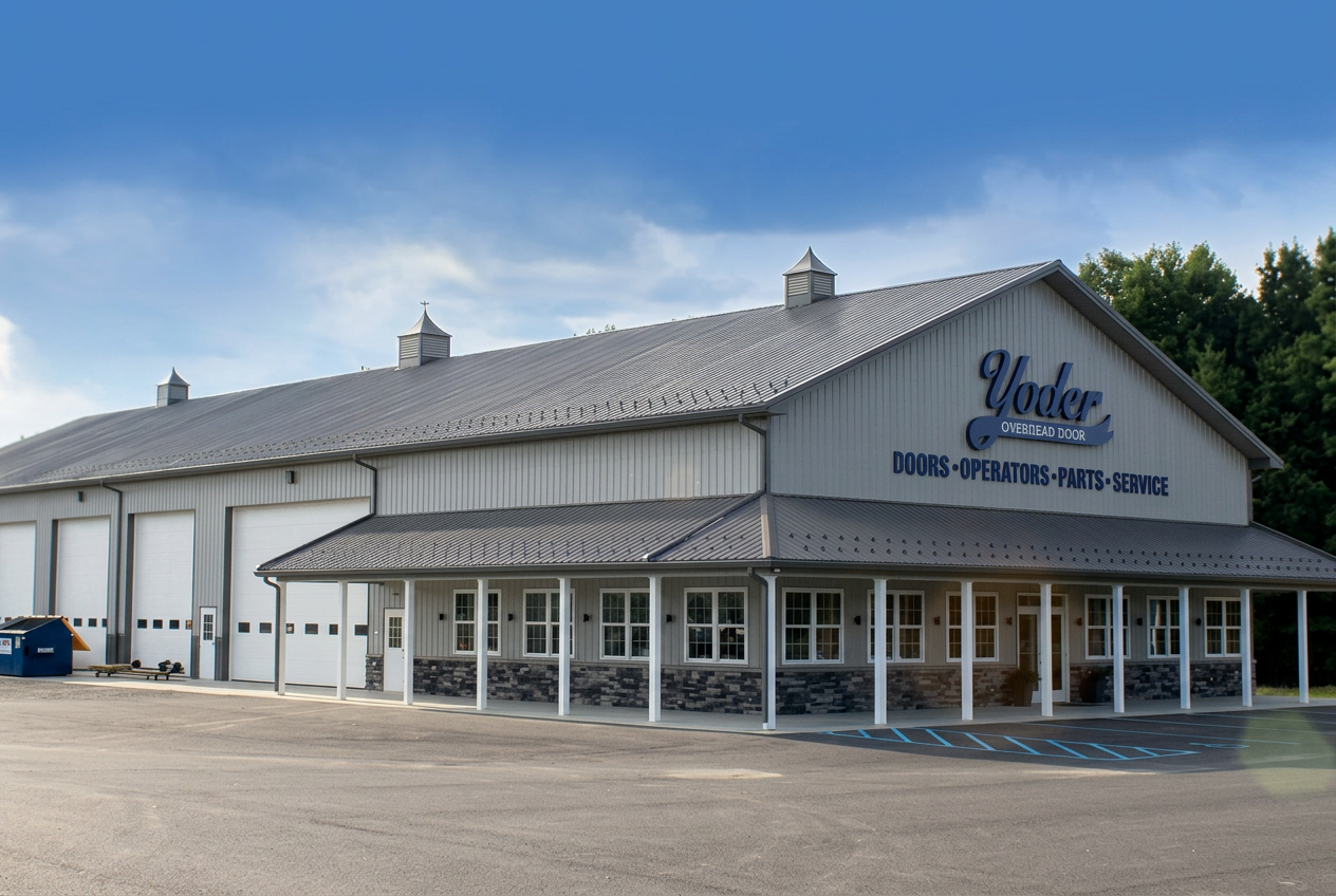 Large industrial building with a gray metal roof and siding, labeled "Yoder Overhead Door," serving as a garage or workshop with multiple large doors, a small parking lot in front, and surrounded by trees under a cloudy sky.