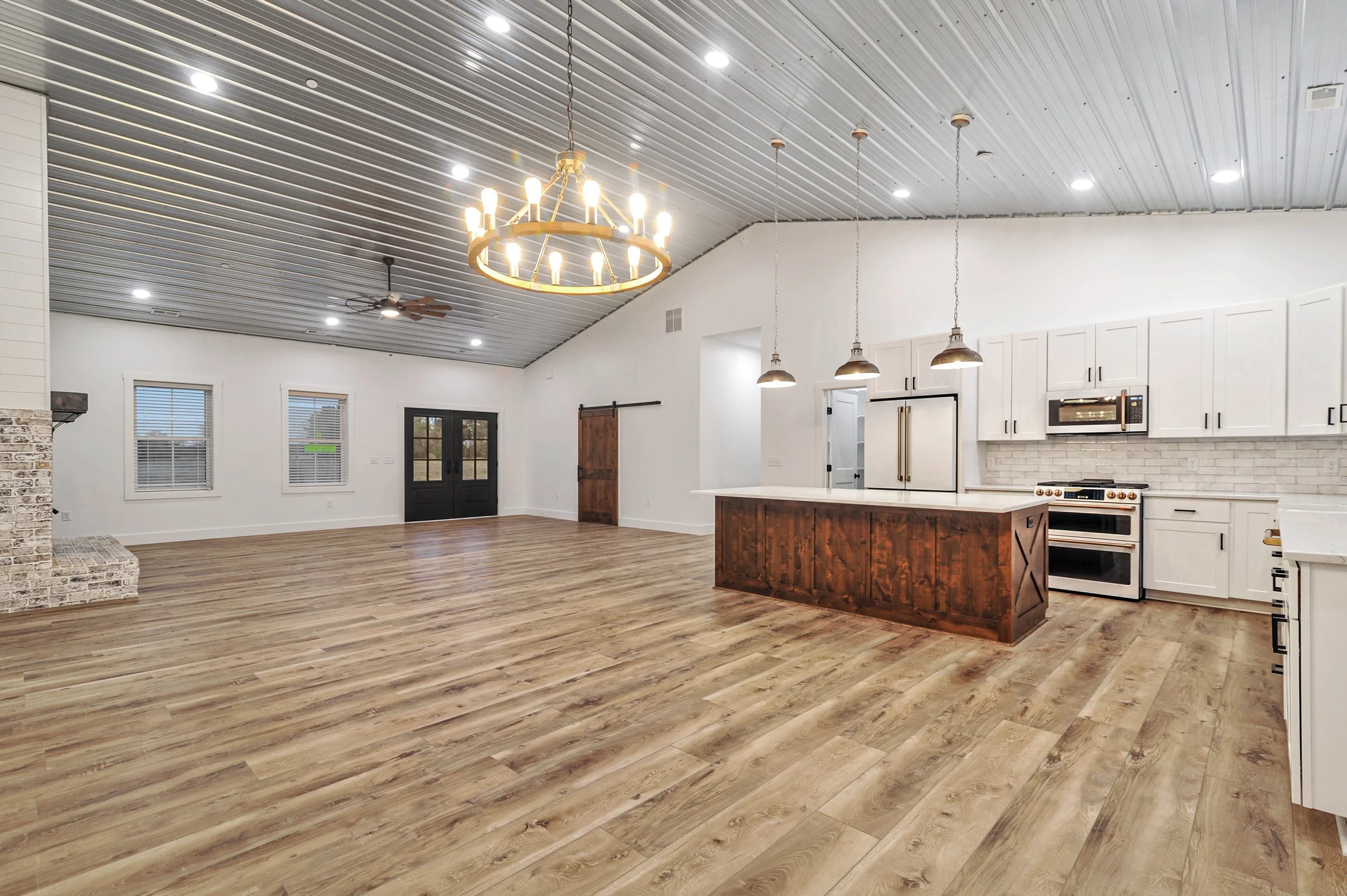 Interior of a spacious, modern kitchen and living area with wooden flooring, white walls, a metal ceiling, and black-framed windows and doors. The kitchen features white cabinets, a brick backsplash, and a central island with a wooden finish. There is a chandelier, pendant lights, and ceiling fans lighting the space.