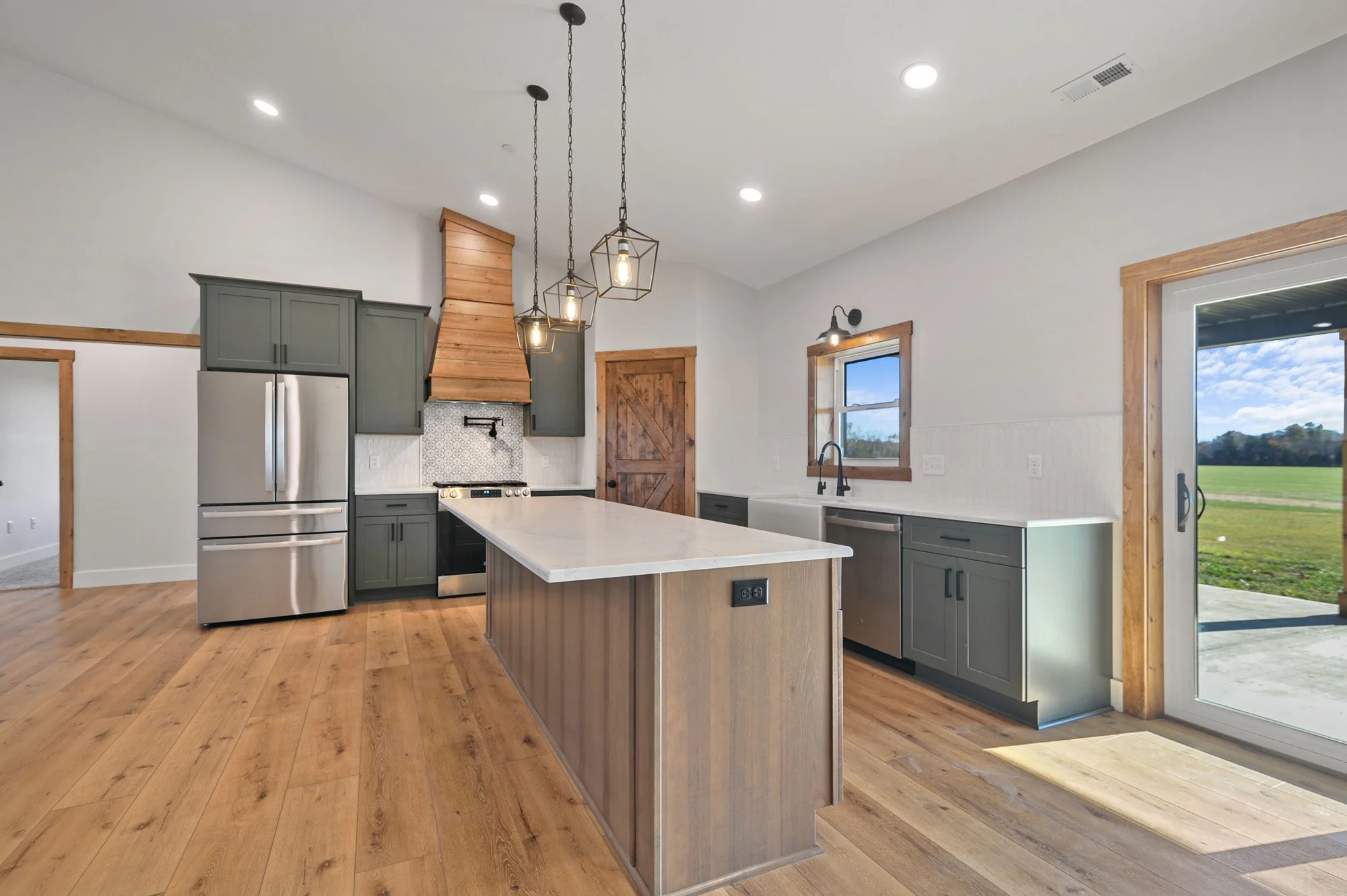 Modern kitchen with gray cabinets, island with white countertop, stainless steel appliances, wooden accents, pendant lights, and large window showing green fields outside.