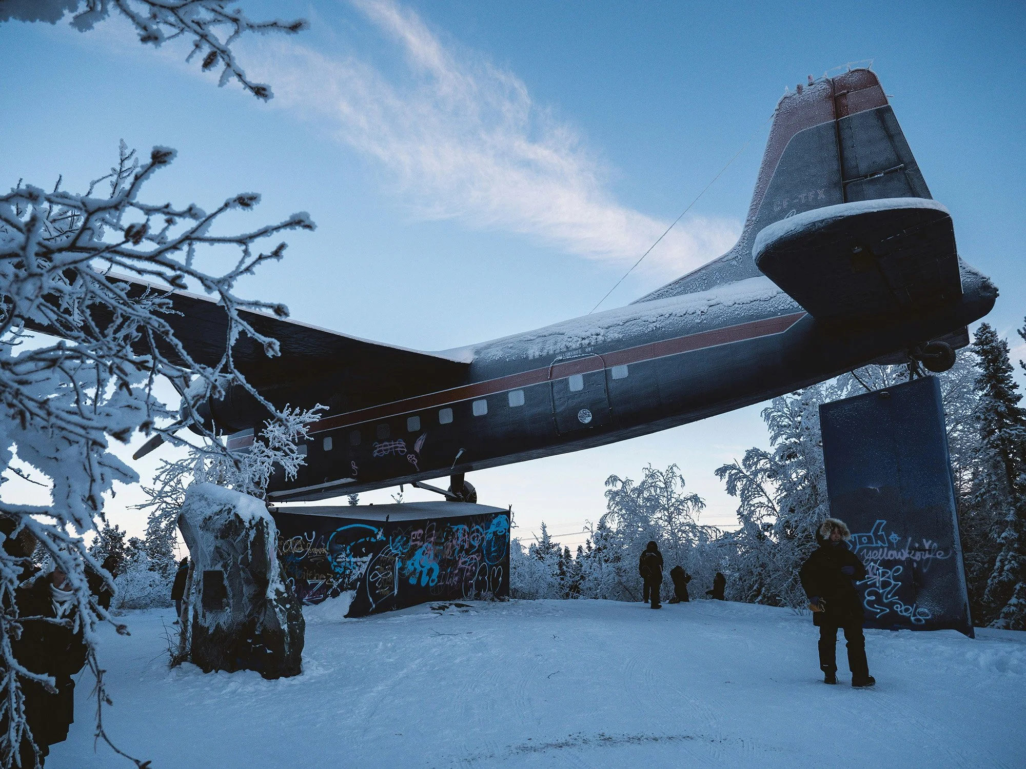 Snow-covered trees and ground with a large helicopter on display, people walking nearby in a snowy outdoor setting under a partly cloudy sky.