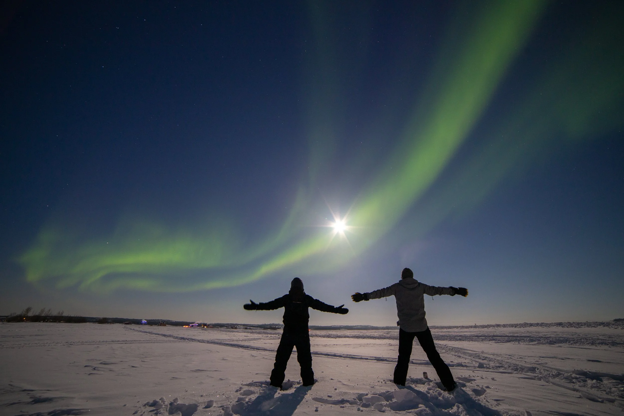 Two people standing in a snowy field at night with their arms raised, watching the Northern Lights in the sky.