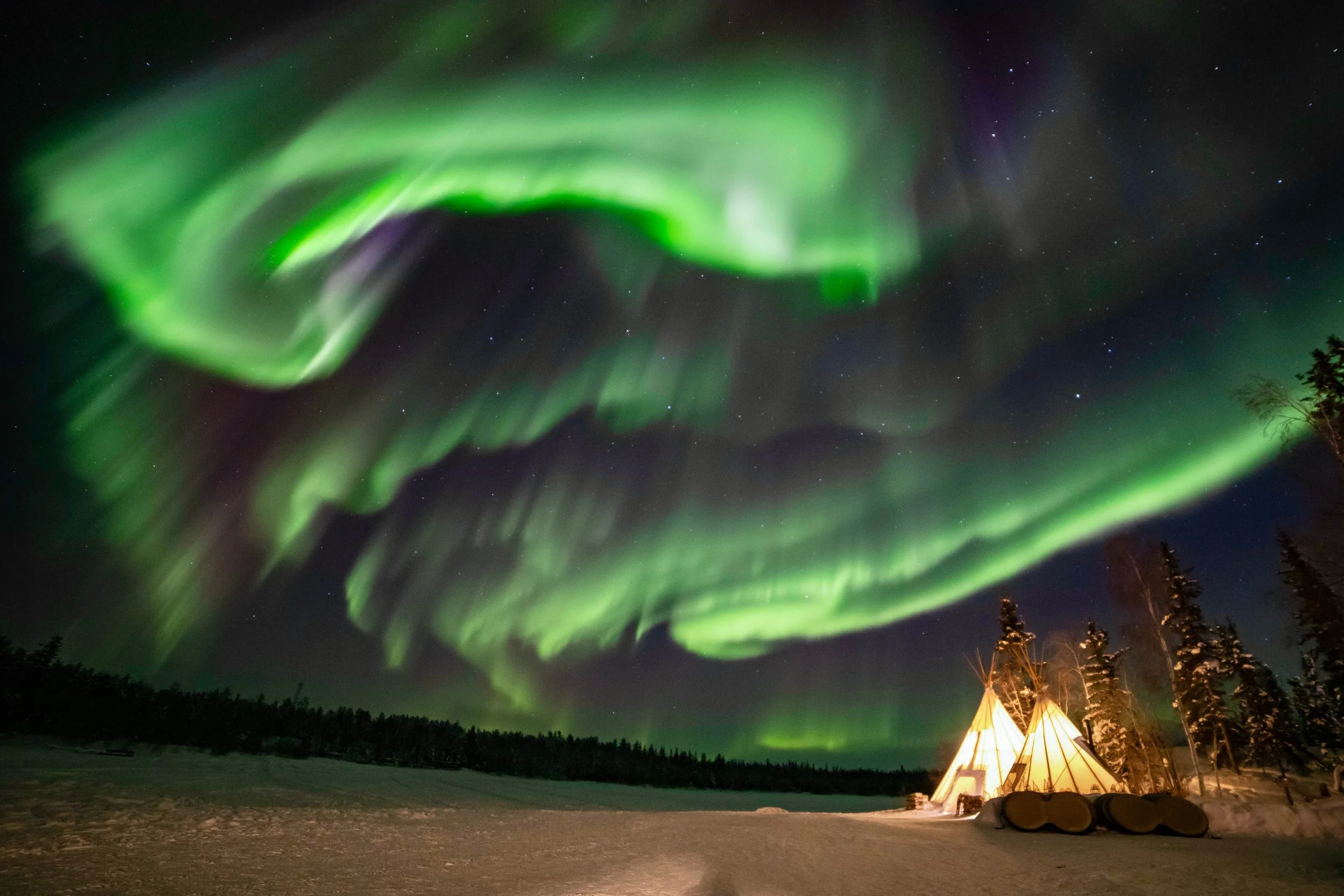 Northern Lights aurora borealis dance across the night sky above a snowy landscape with trees and a tipi tent glowing softly.