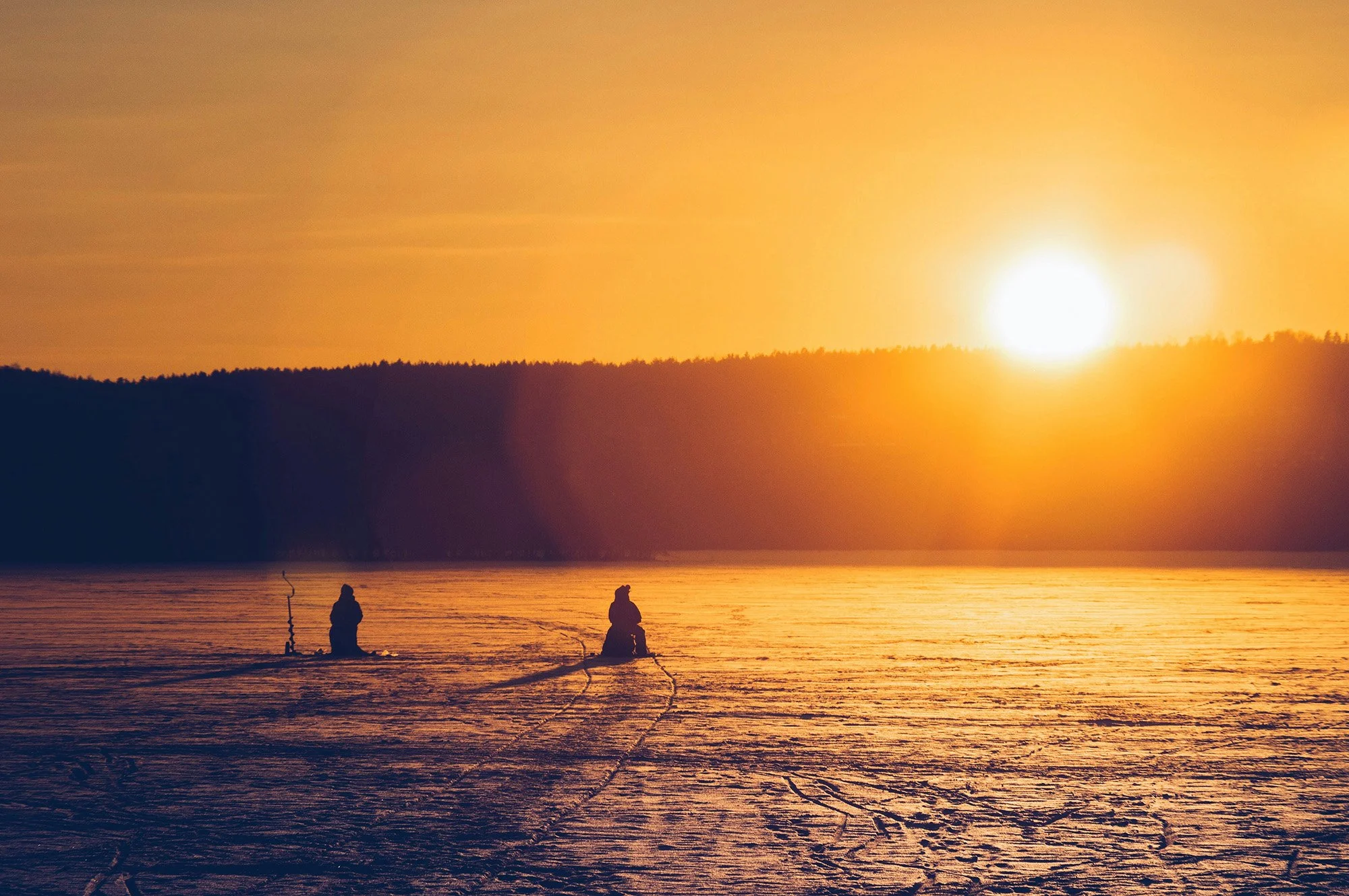 Silhouettes of two people fishing by the water at sunset, with a large glowing sun low on the horizon and a distant tree-covered hillside in the background.
