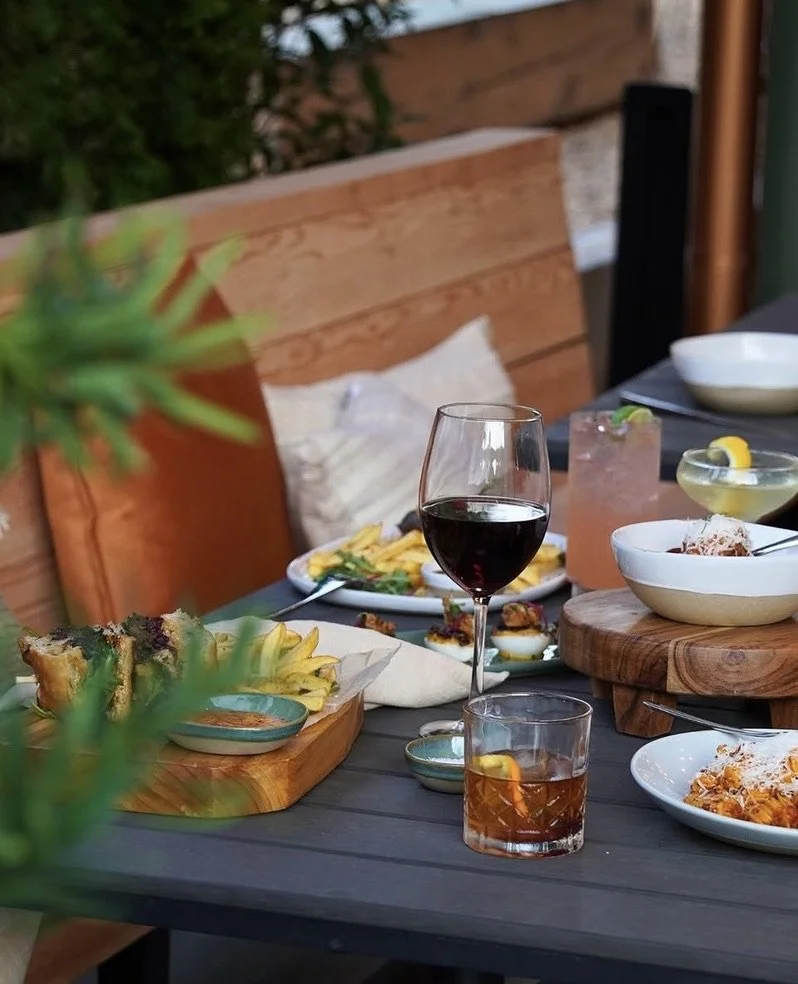Outdoor dining table with various food and drinks, including a glass of red wine, a pink cocktail, a glass of whiskey, plates of sushi, fries, and appetizers, with a wooden bench and greenery in the background.