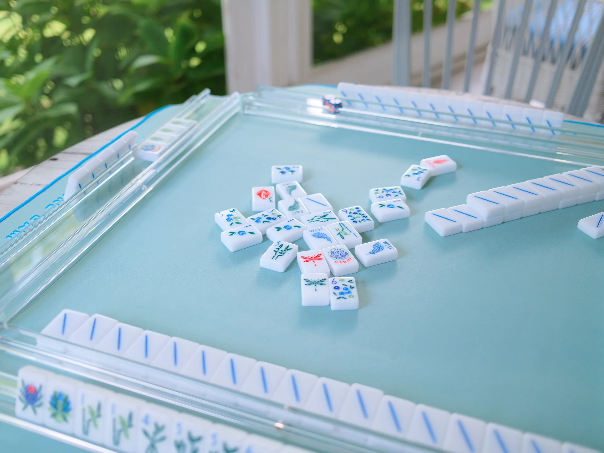 A close-up of The Mahjong Line tile game setup on a light blue mat, with several tiles arranged in a row and some scattered around, against a green leafy background.