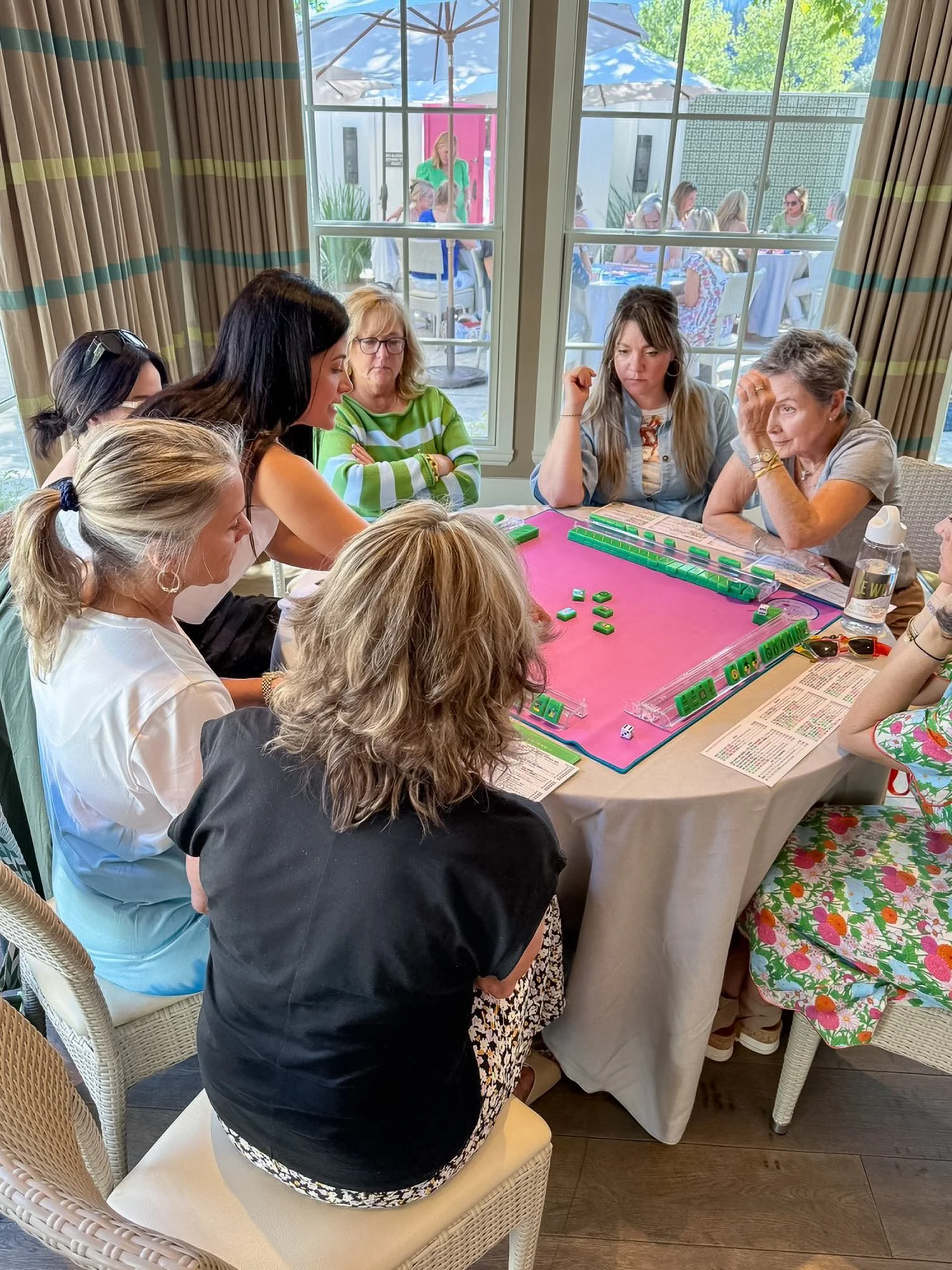 Group of women gathered around a table playing a game of Mahjong with green tiles and dice at a table near large windows showing an outdoor patio with umbrellas and other people seated at tables.