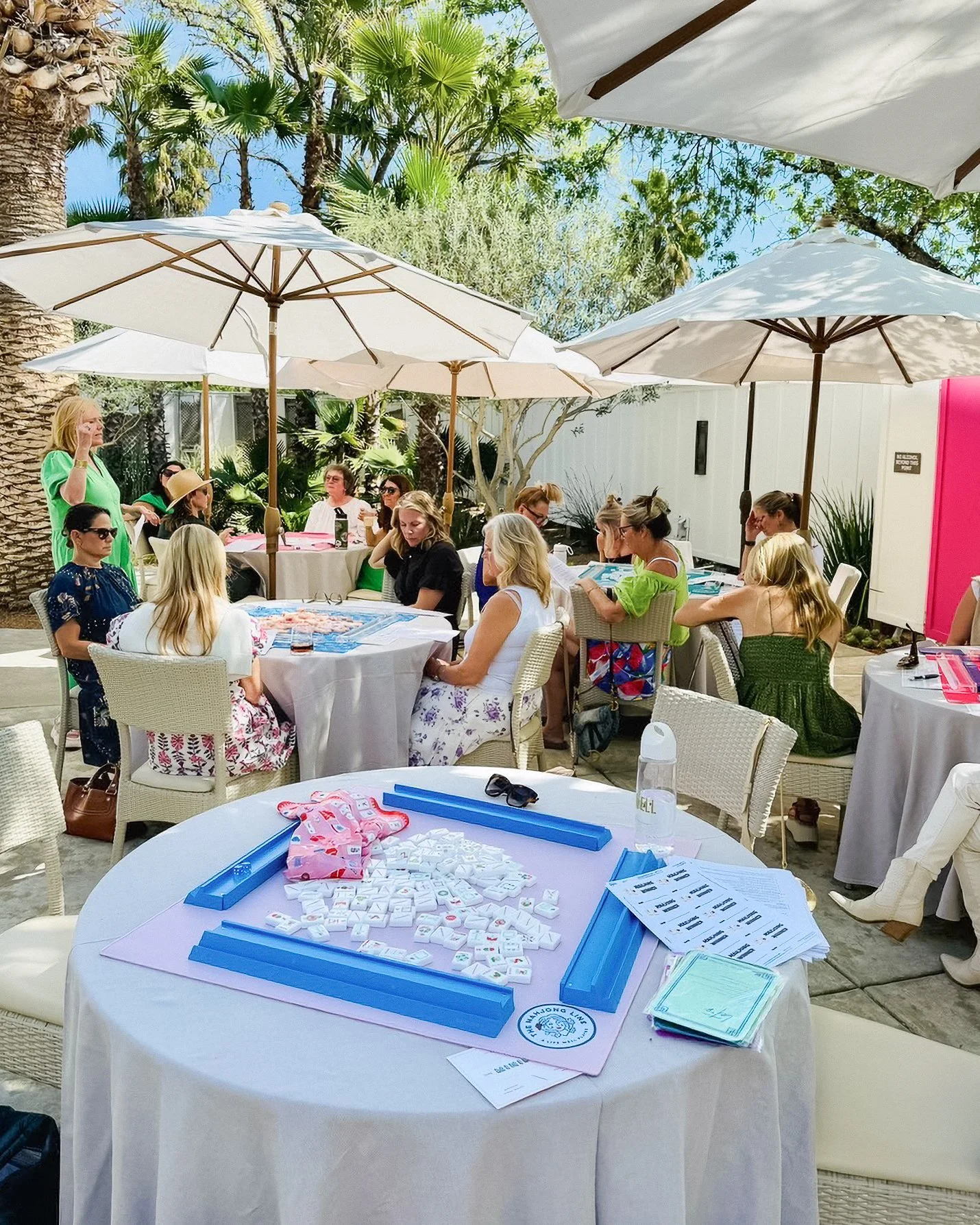 Outdoor gathering with white tables, chairs, and umbrellas, where women are playing mahjong with tiles and score sheets, under a sunny sky with trees in the background.