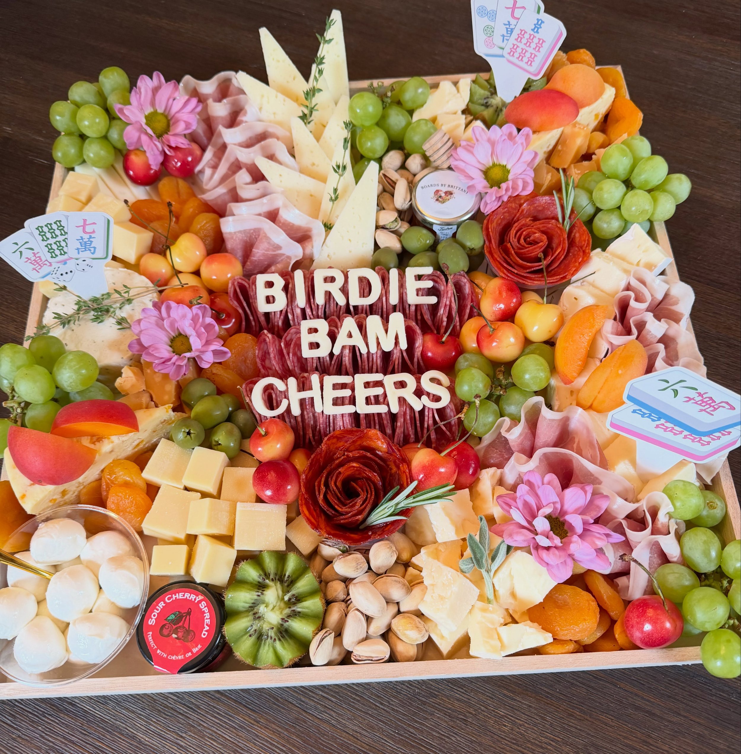 A wooden charcuterie tray filled with cheese, grapes, cherries, sliced peaches, pistachios, and flowers. The cheese spells "BIRDIE," "BAM," and "CHEERS."