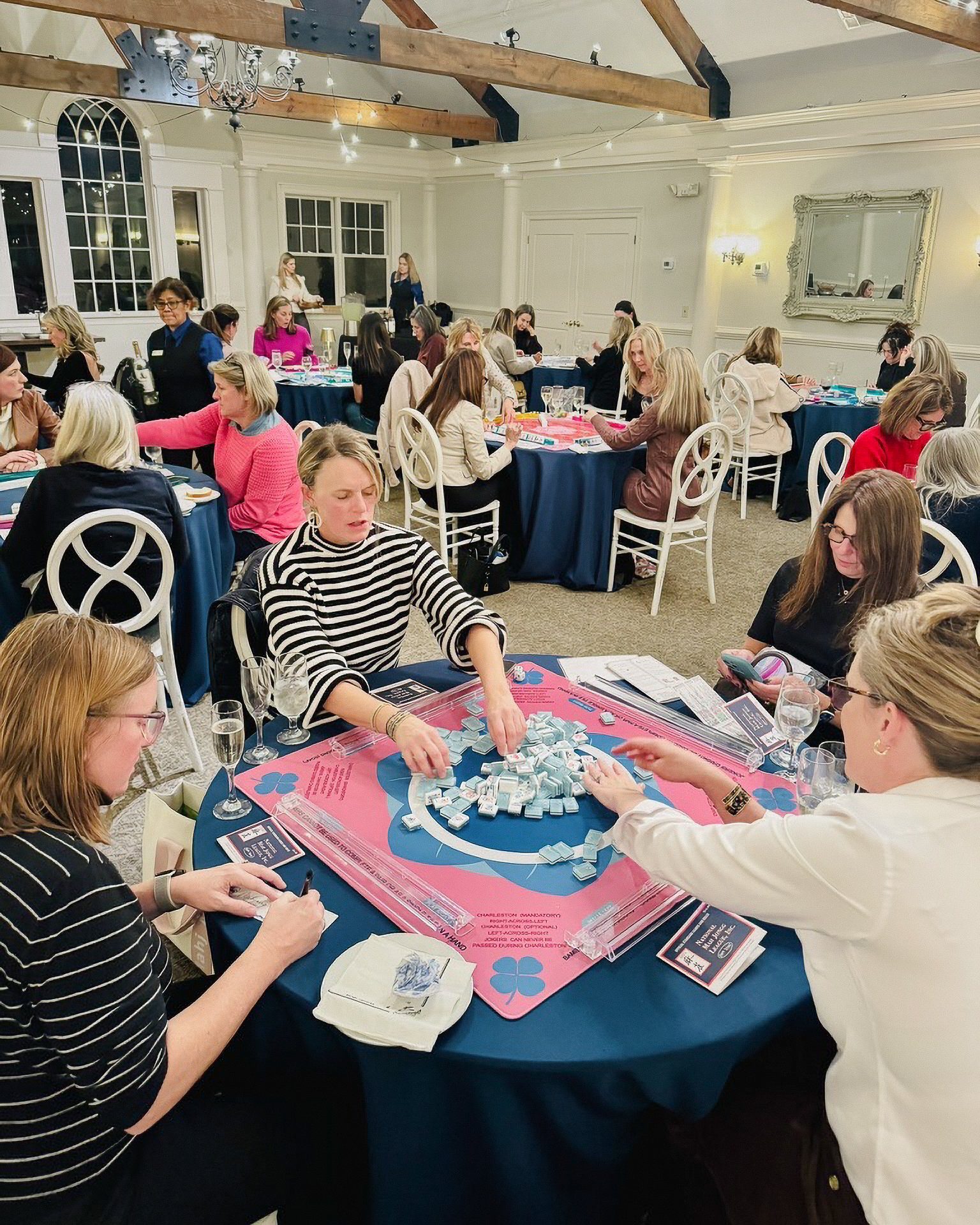 Women playing Mahjong at a dinner event, with a woman in white and a woman in a red sweater visible, and large windows in the background.