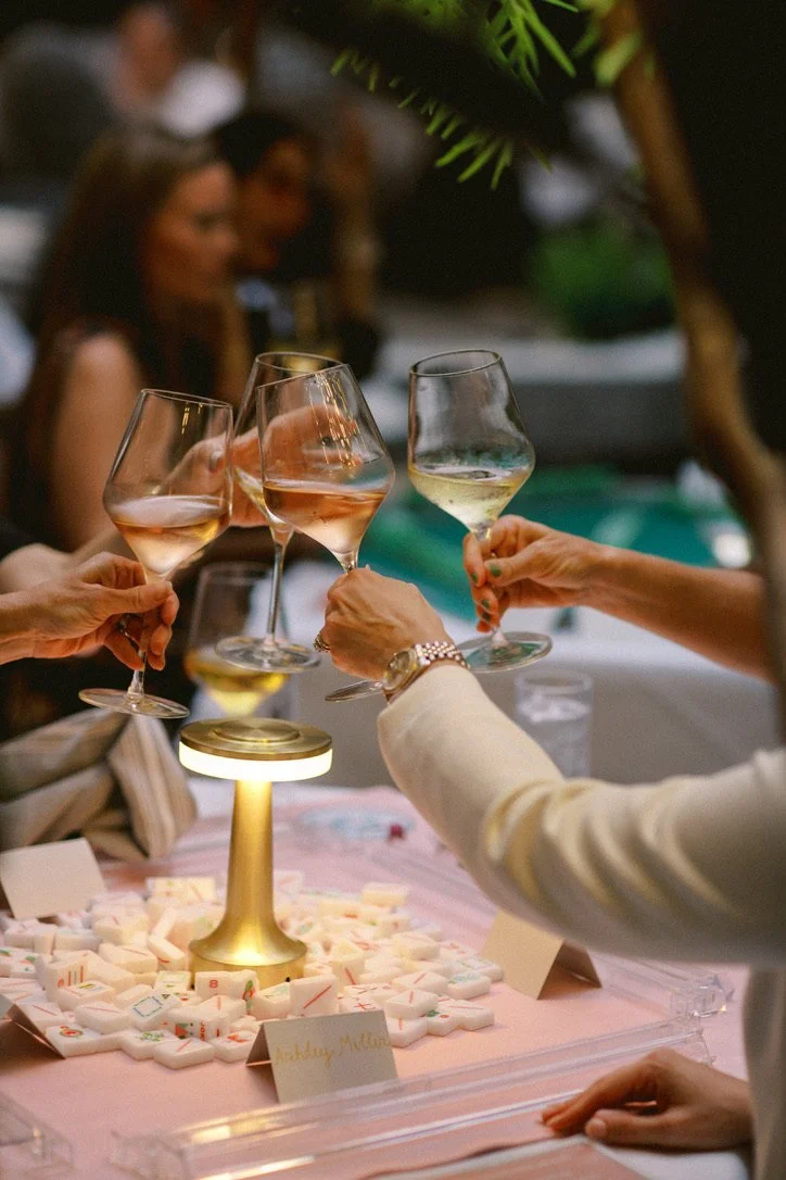 People raising glasses of wine in a toast at a celebration table decorated with Mahjong tiles and a small illuminated lamp, with blurred guests in the background.