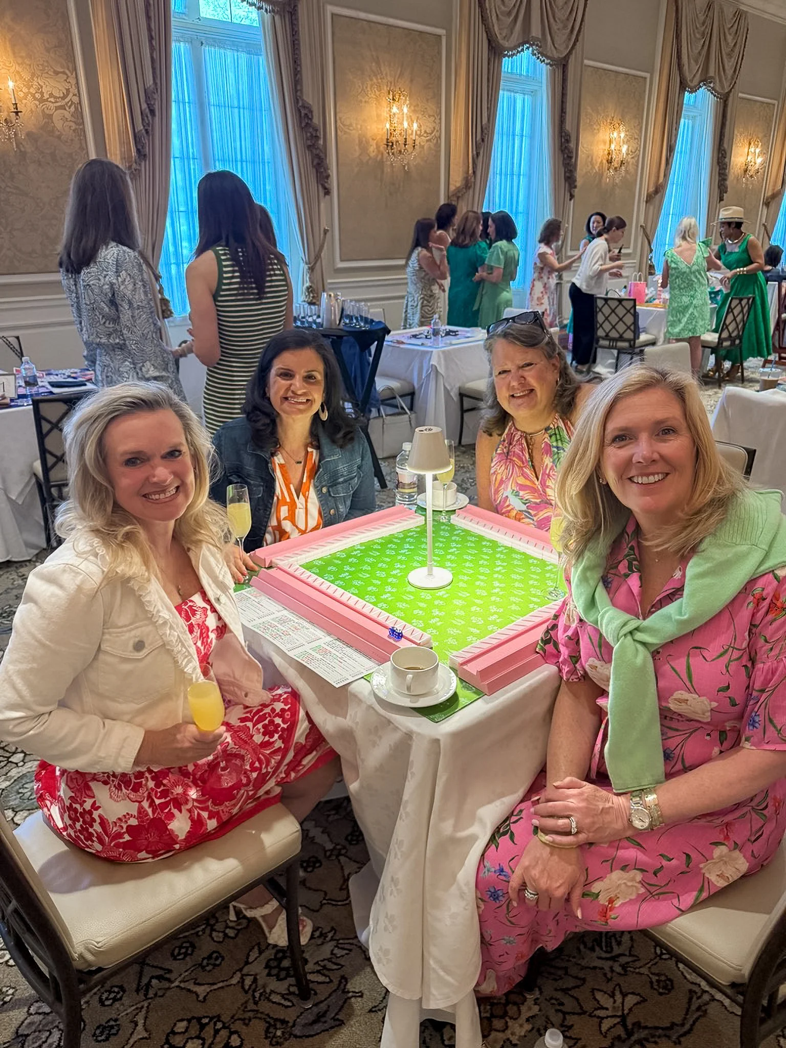 A group of women at a charitable gathering playing Mahjong. They are seated around a table with drinks, and some women are standing in the background talking. A decorated banquet room with tall windows, drapery, and chandelier lighting.