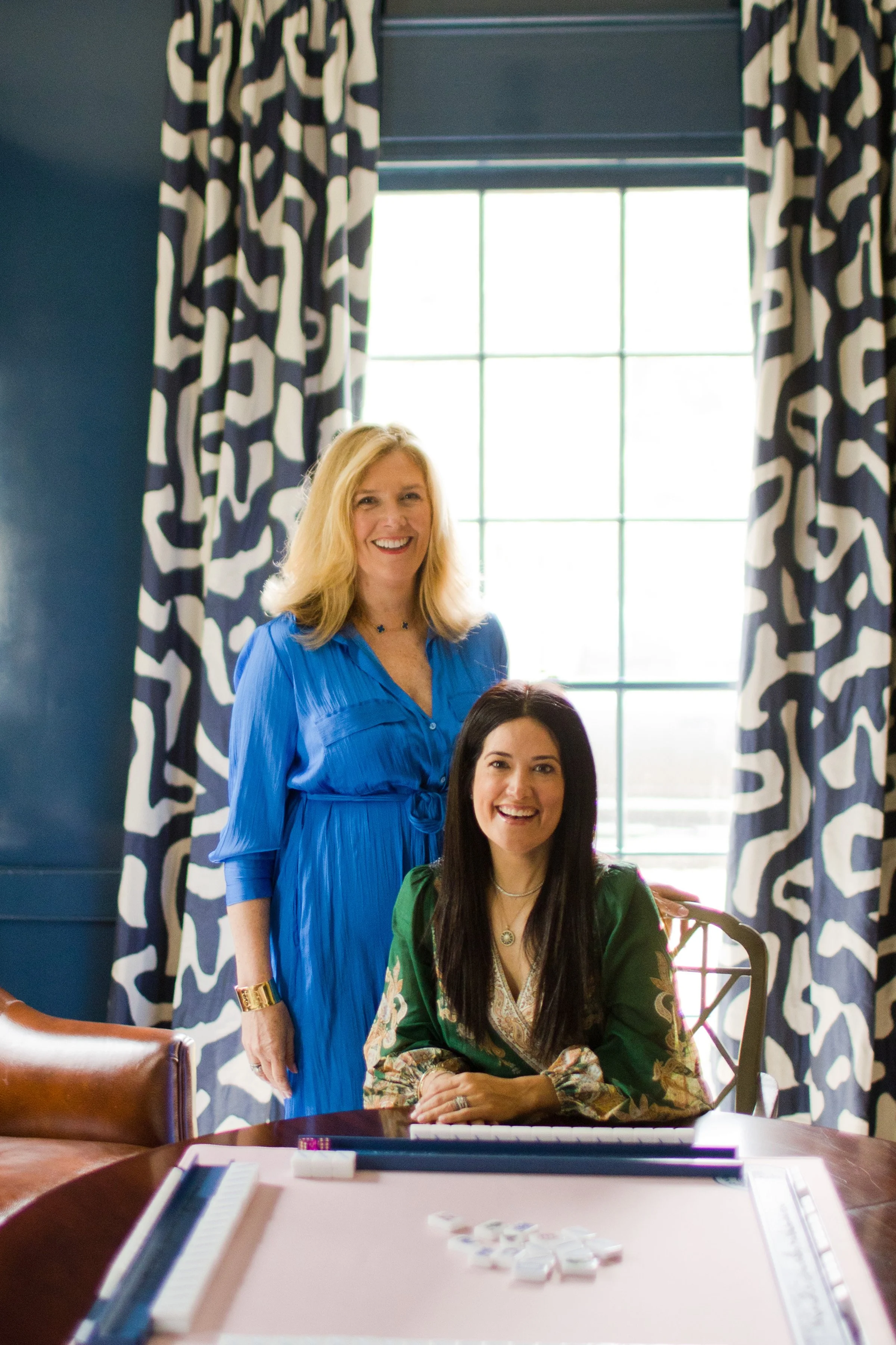 Two women in a room, one standing and smiling, the other sitting at a table with a game of Mahjong, with large windows and patterned curtains in the background.
