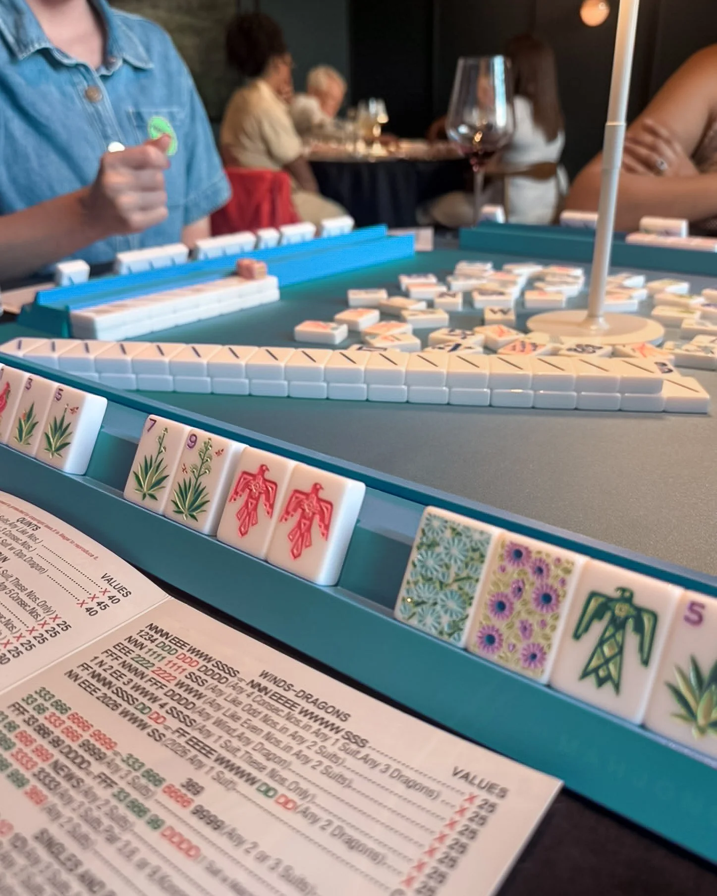 Close-up of a Mahjong game table with tiles, a person holding a tile, and a score sheet in the foreground. In the background, guests are seated at tables playing Mahjong.