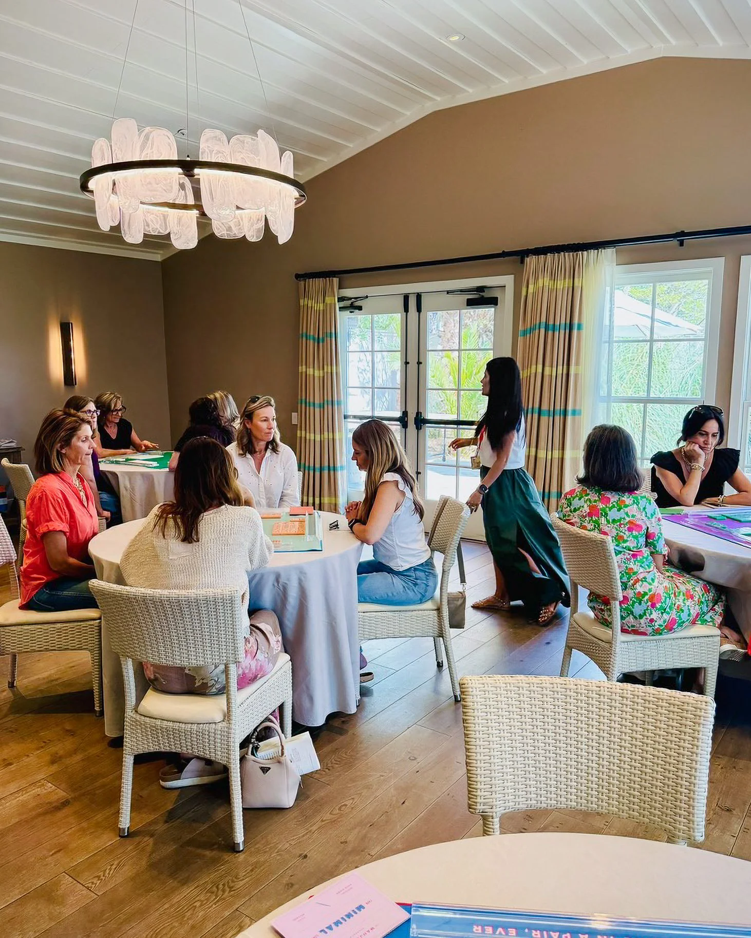 A group of women sitting around a table in a well-lit room with large windows, listening to a standing woman who appears to be giving a presentation or leading a discussion.