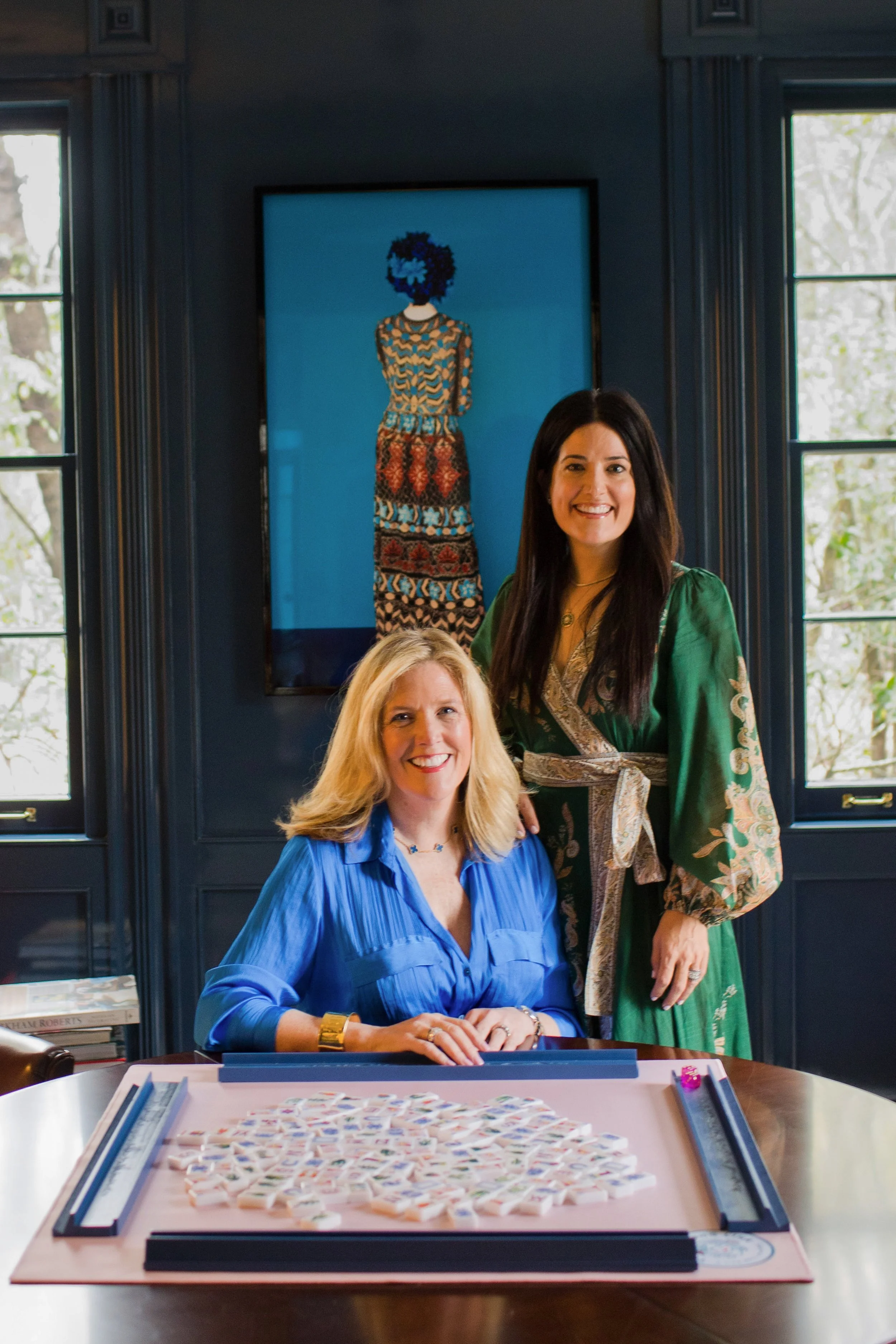 Liz Liu and Isabel Rice, co-founders of Southern Mahjong Co., in a room with a large window, a framed piece of art behind them, and a Mahjong board on the table with tiles arranged on it.