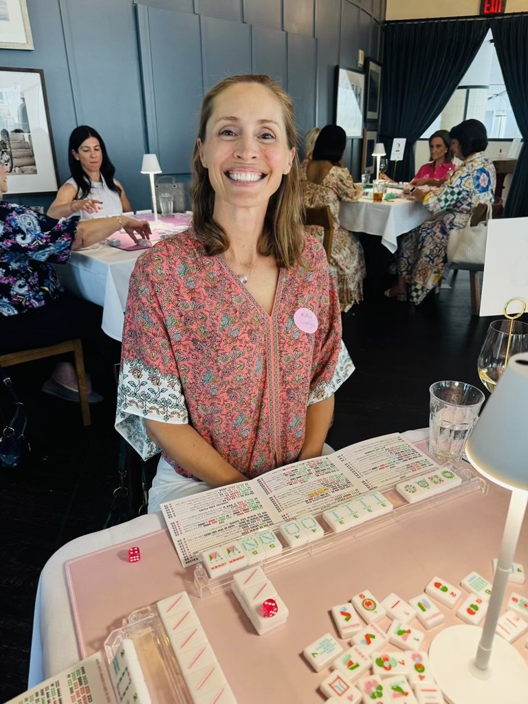 A woman with shoulder-length light brown hair smiling at the camera is sitting at a table playing mahjong, with mahjong tiles and scorecards in front of her, in a well-lit indoor space with other women sitting at tables in the background.
