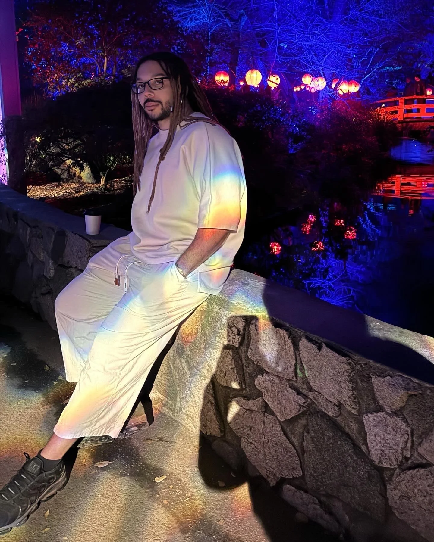 A person with long dreadlocks, glasses, and a white outfit sitting on a stone bench at night with colorful lanterns and illuminated trees in the background.