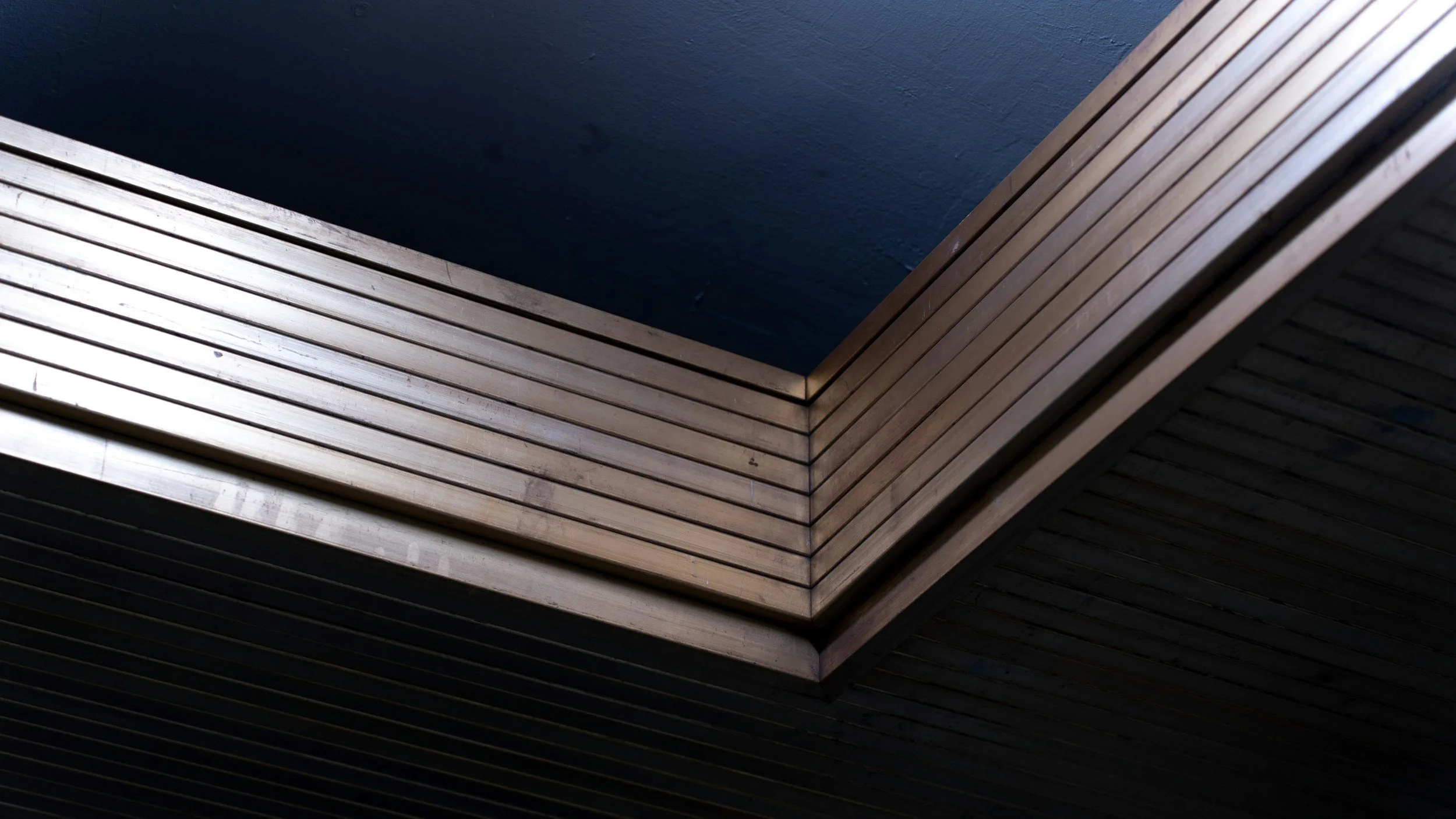 A modern architectural ceiling structure featuring layered wooden slats forming a geometric pattern with a rectangular opening in the center, contrasting with a dark background.