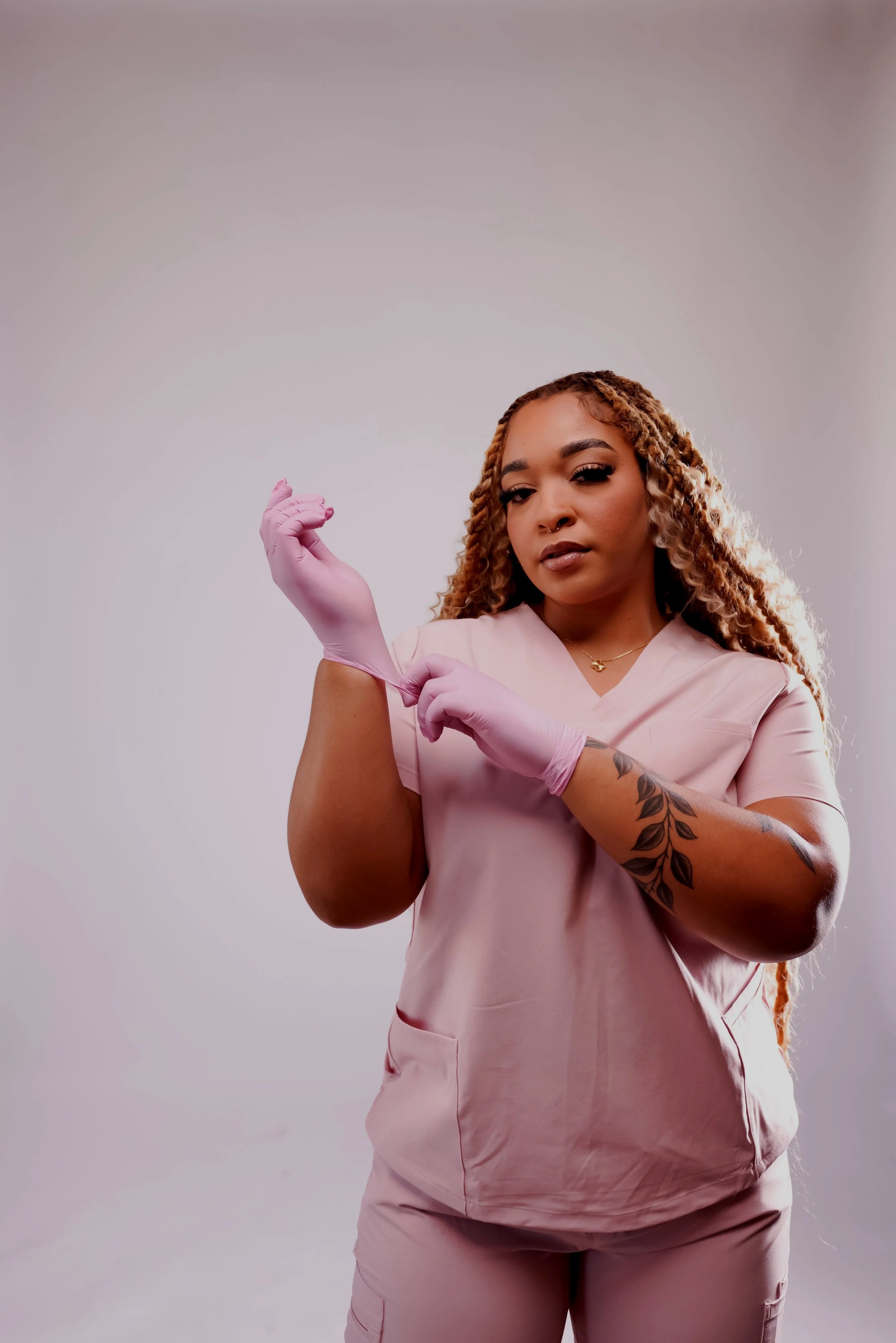 A woman with curly hair in scrubs and a tattoo on her arm is putting on pink gloves against a plain light gray background.