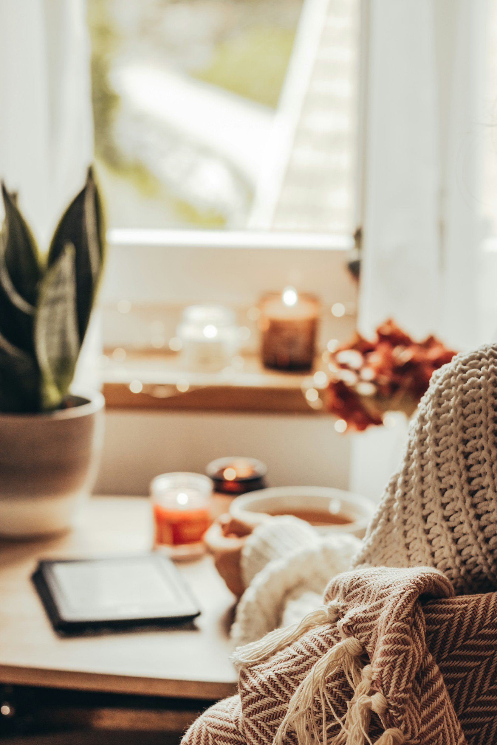 A cozy indoor scene with a knitted blanket, a cup of coffee, candles, a plant, and a notebook on a wooden table near a window with sunlight.