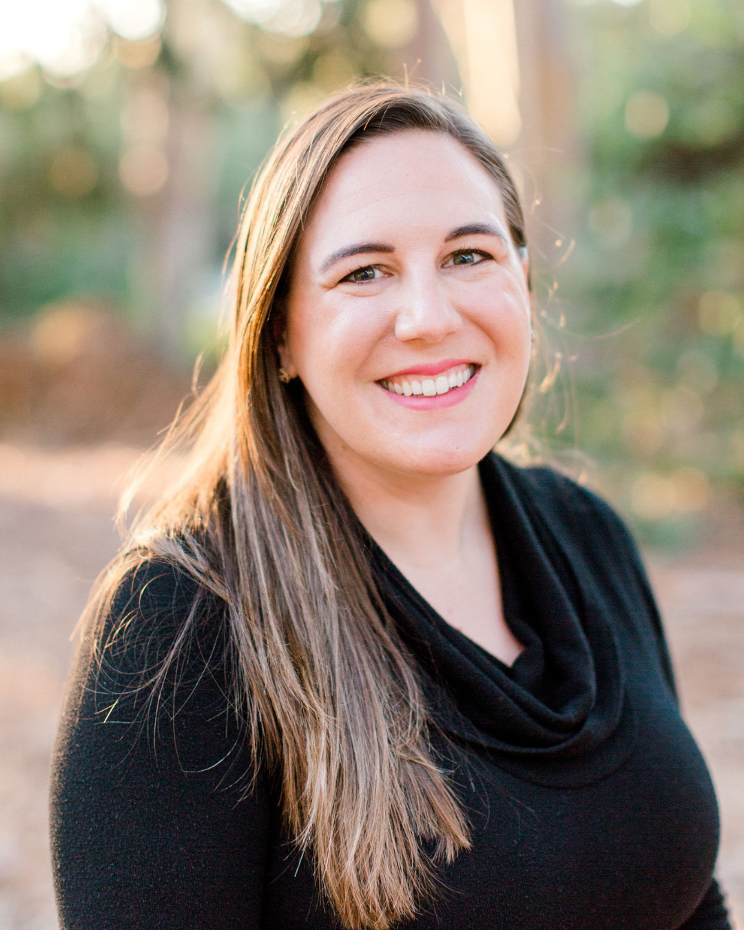 A smiling woman with long brown hair wearing a black top outdoors, with trees in the background