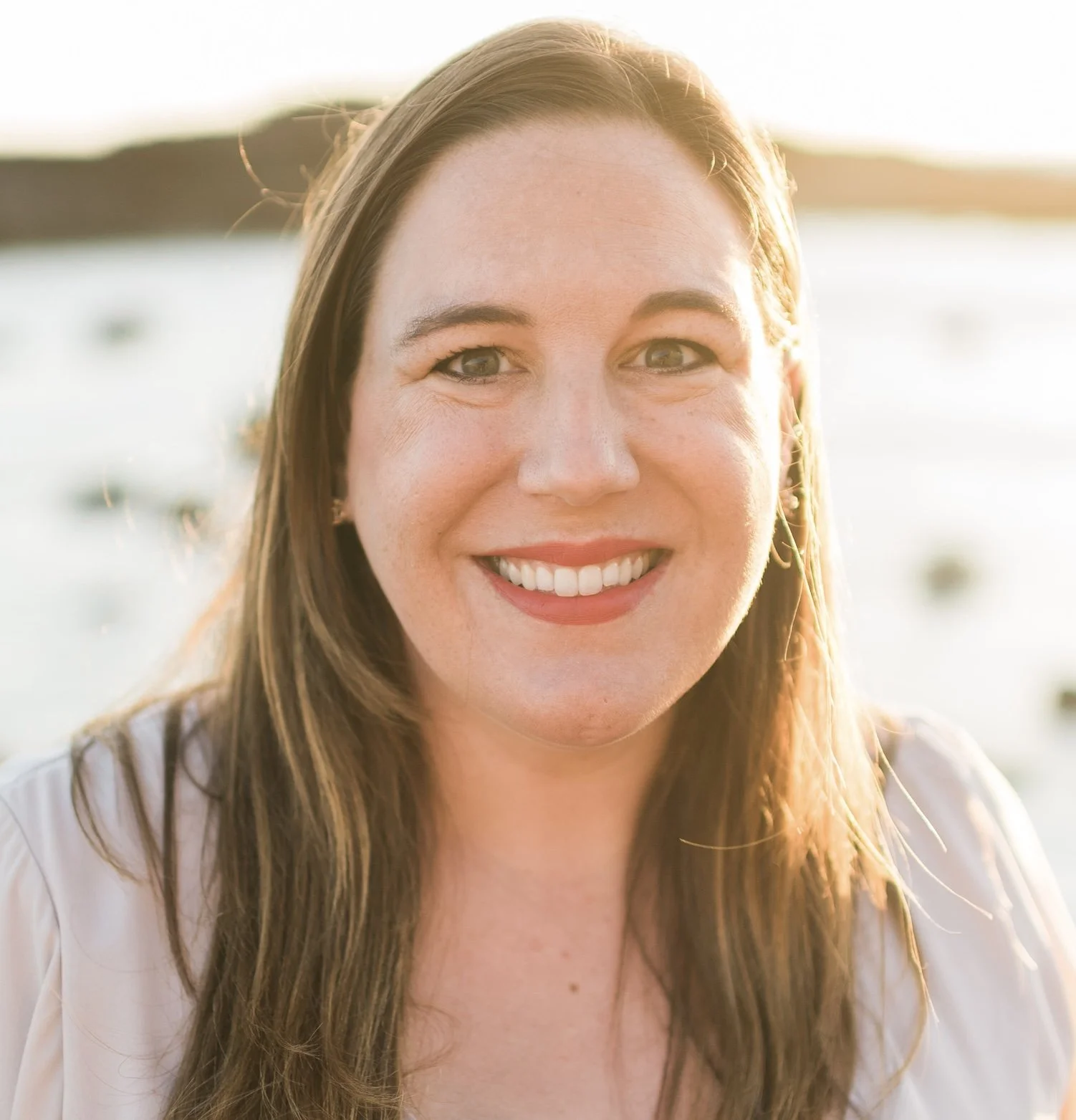 Close-up portrait of a woman smiling with long brown hair, sunlight backlighting her, blurred outdoor background.