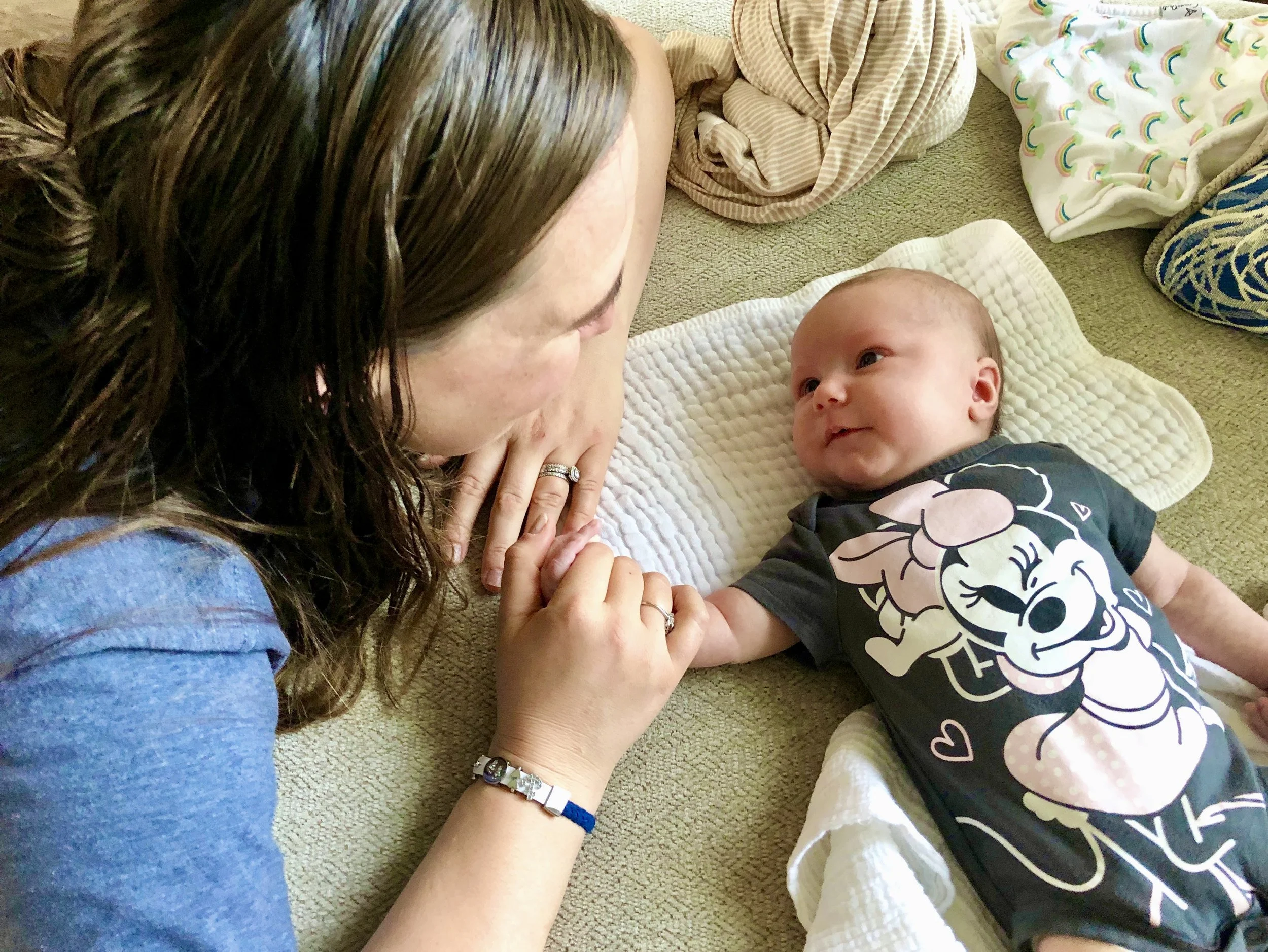 A woman with long brown hair and a bracelet on her wrist lies on her stomach looking at a baby who is lying on a white quilt on the floor. The baby is wearing a black shirt with a Minnie Mouse design and is looking at the woman with a smile. There are clothes and blankets with rainbow and rainbow-like patterns around them.